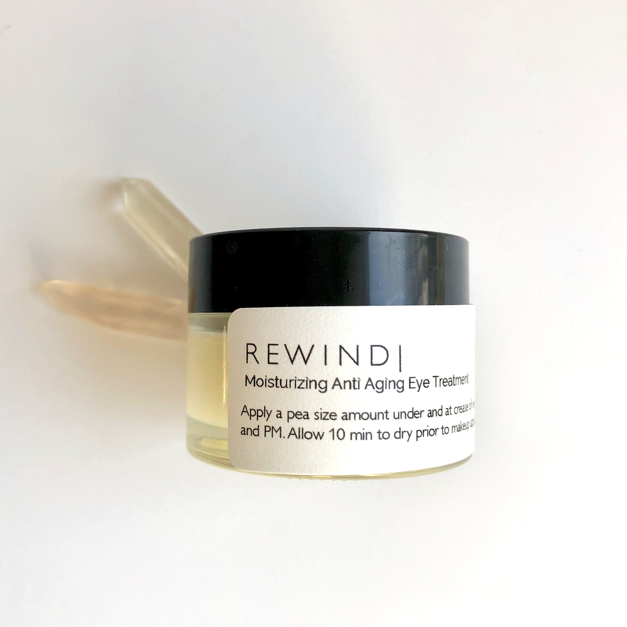 Eye treatment jar with black lid, clear glass, and white label with text REWINDI