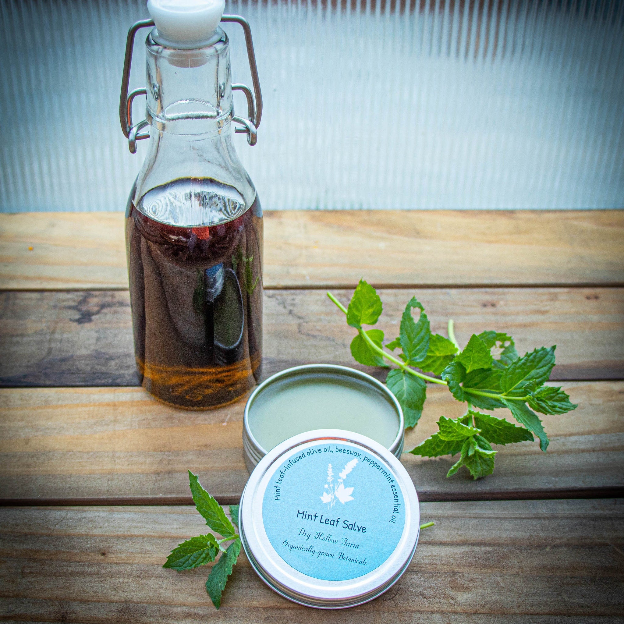 Mint salve in open metal tin, with green leaf, and glass bottle on wooden surface.