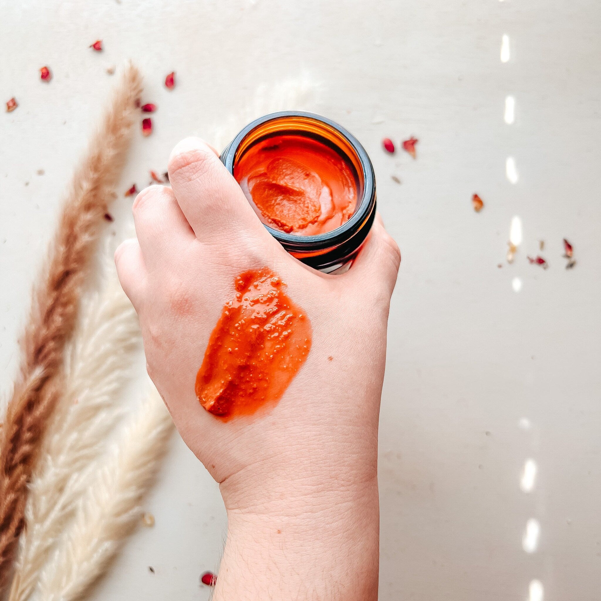 Face mask jar held in hand with orange paste, brown glass jar, and white background