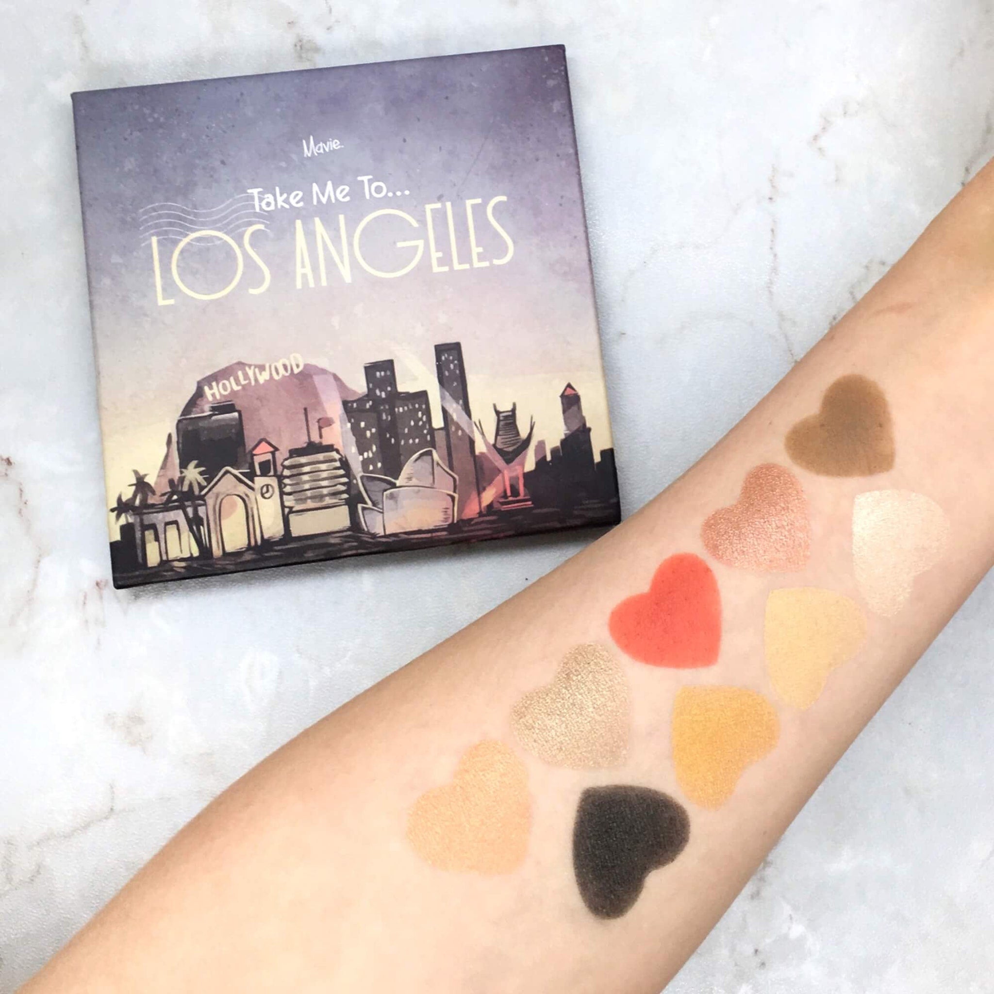 Eyeshadow palette with "Take Me To Los Angeles" text, heart-shaped swatches, and various colors on arm