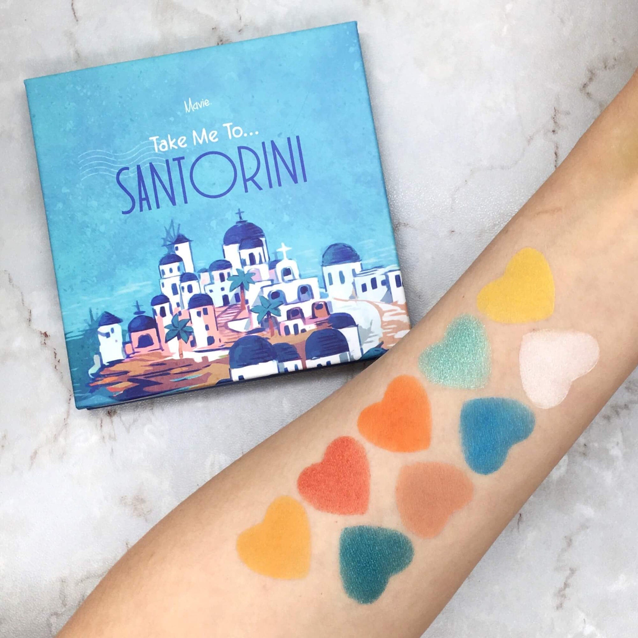 Eyeshadow palette with Mavie Cosmetics branding, blue and white packaging, and heart-shaped swatches on arm