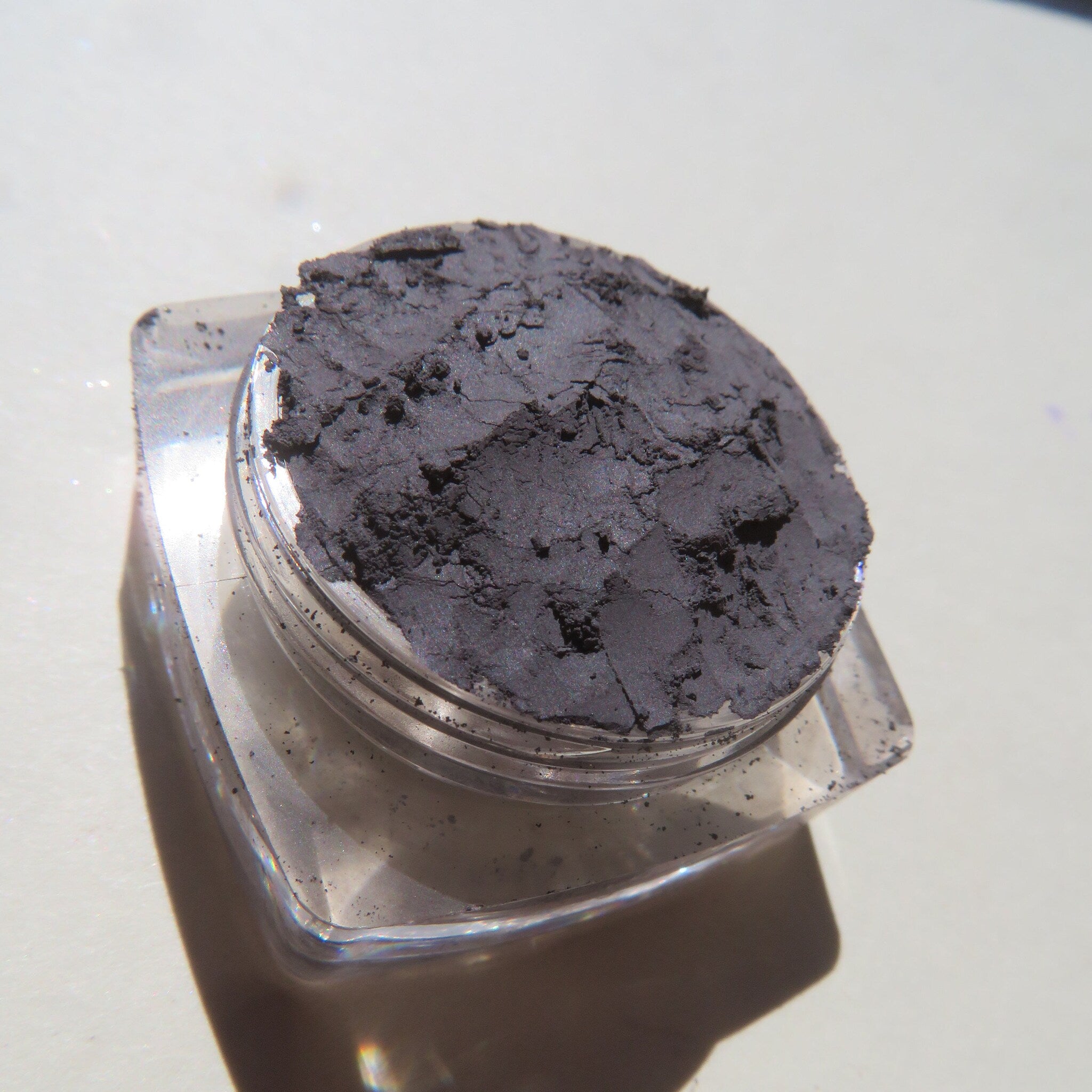 Eyeshadow, dark gray powder, clear plastic jar, textured surface, close-up view