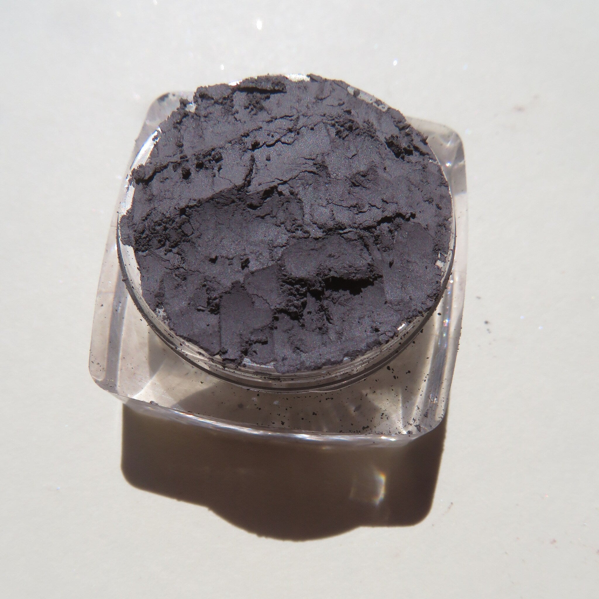 Eyeshadow in dark gray powder with textured appearance in clear plastic container