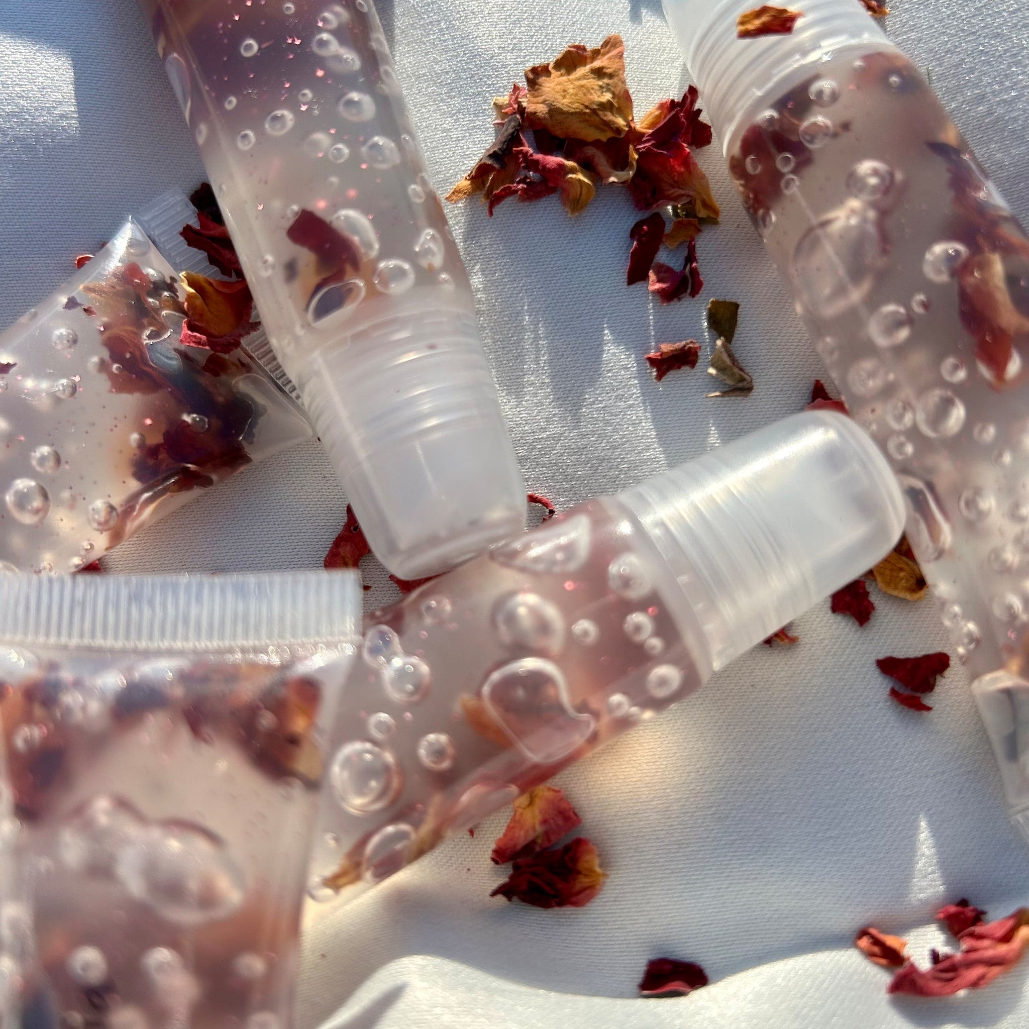 Lip gloss tubes with clear liquid, rose petals, and bubbles on white fabric