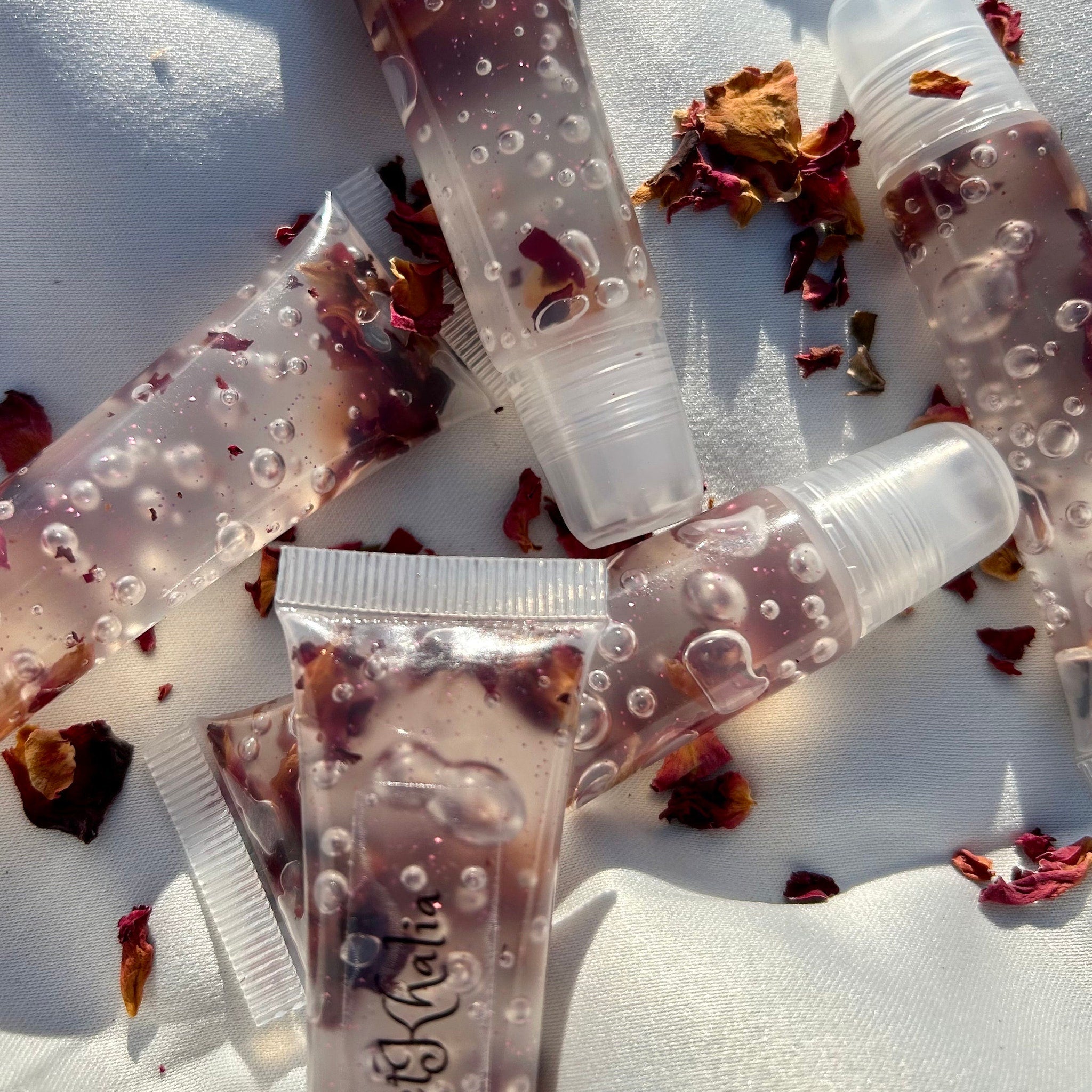Lip gloss tubes with clear liquid, rose petals, and "Khalia" brand text visible on white fabric