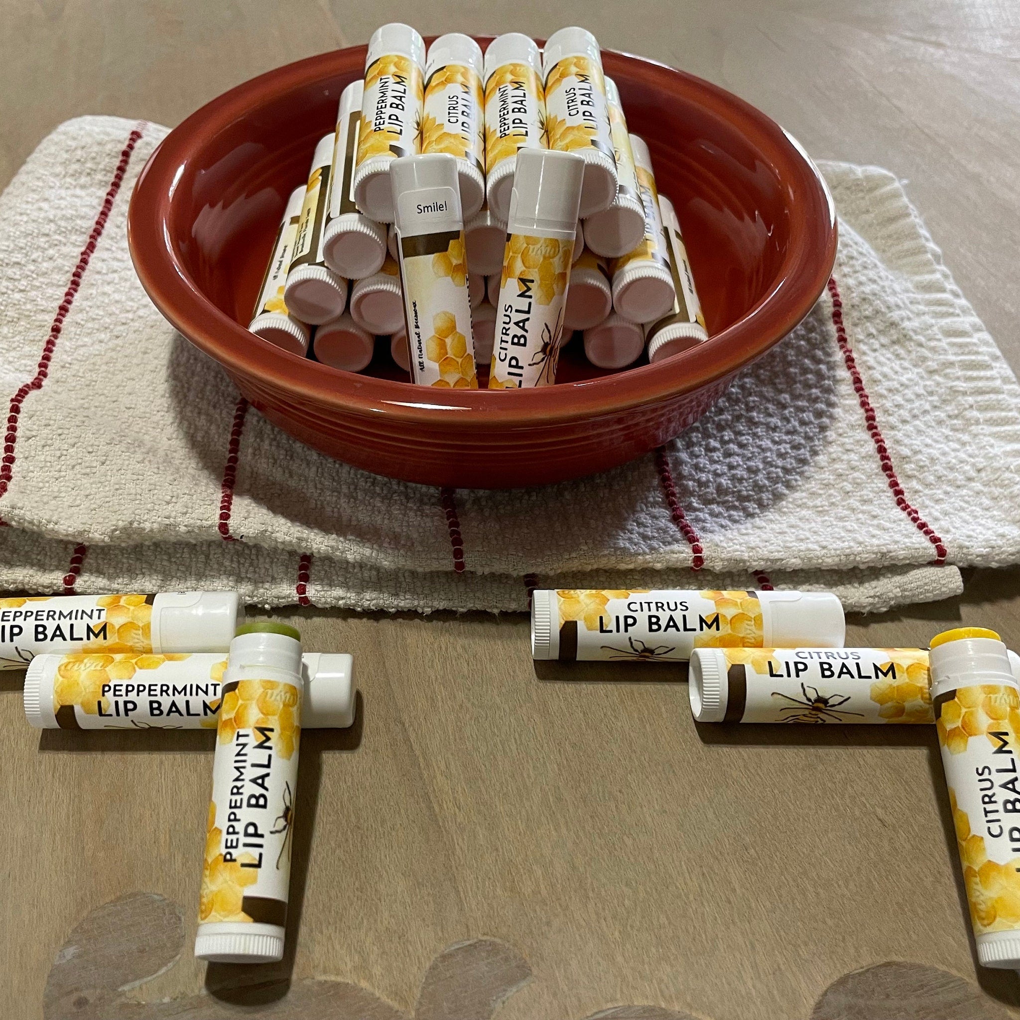 Lip balm tubes with white, yellow, and brown labels, stacked in a red bowl on a white cloth