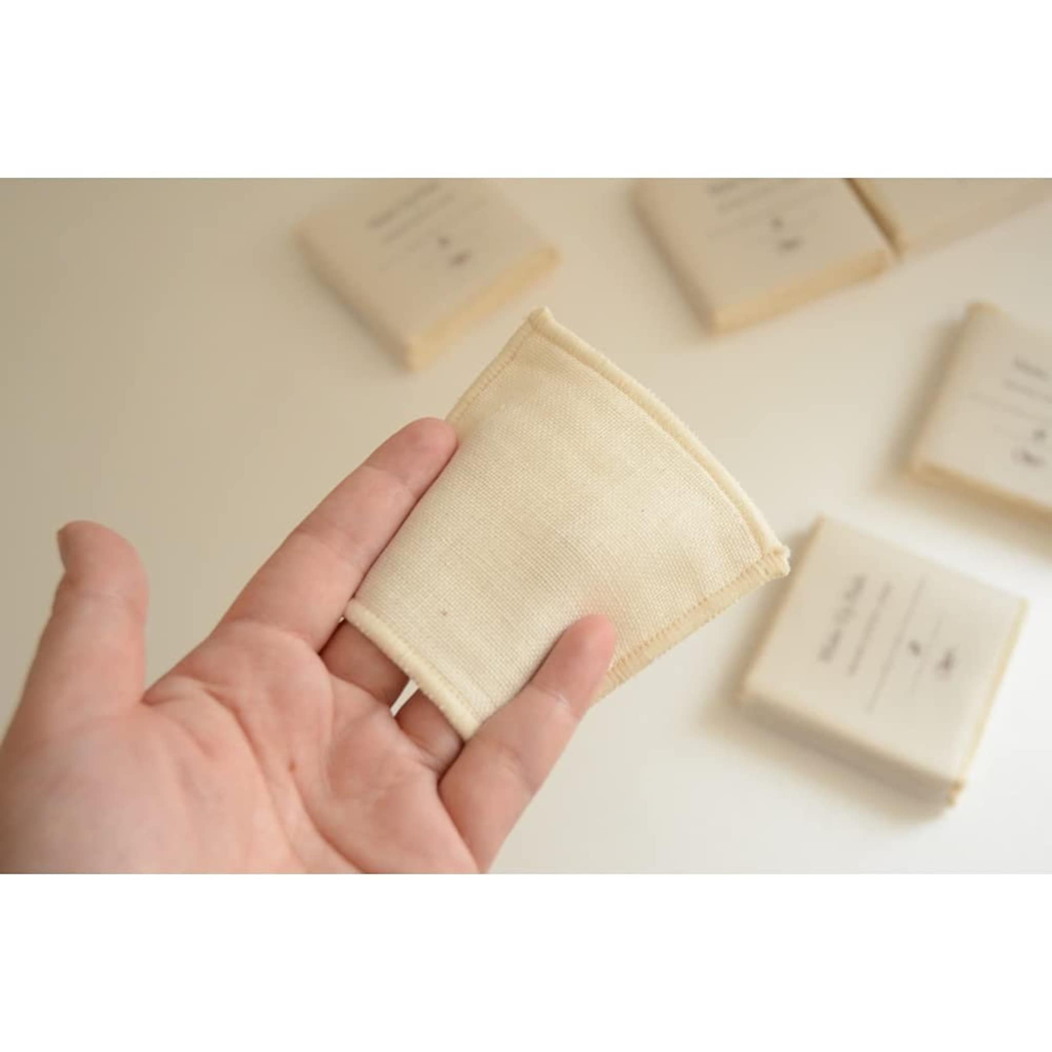 Reusable cotton pad, cream fabric, square shape, held in hand with other pads in background