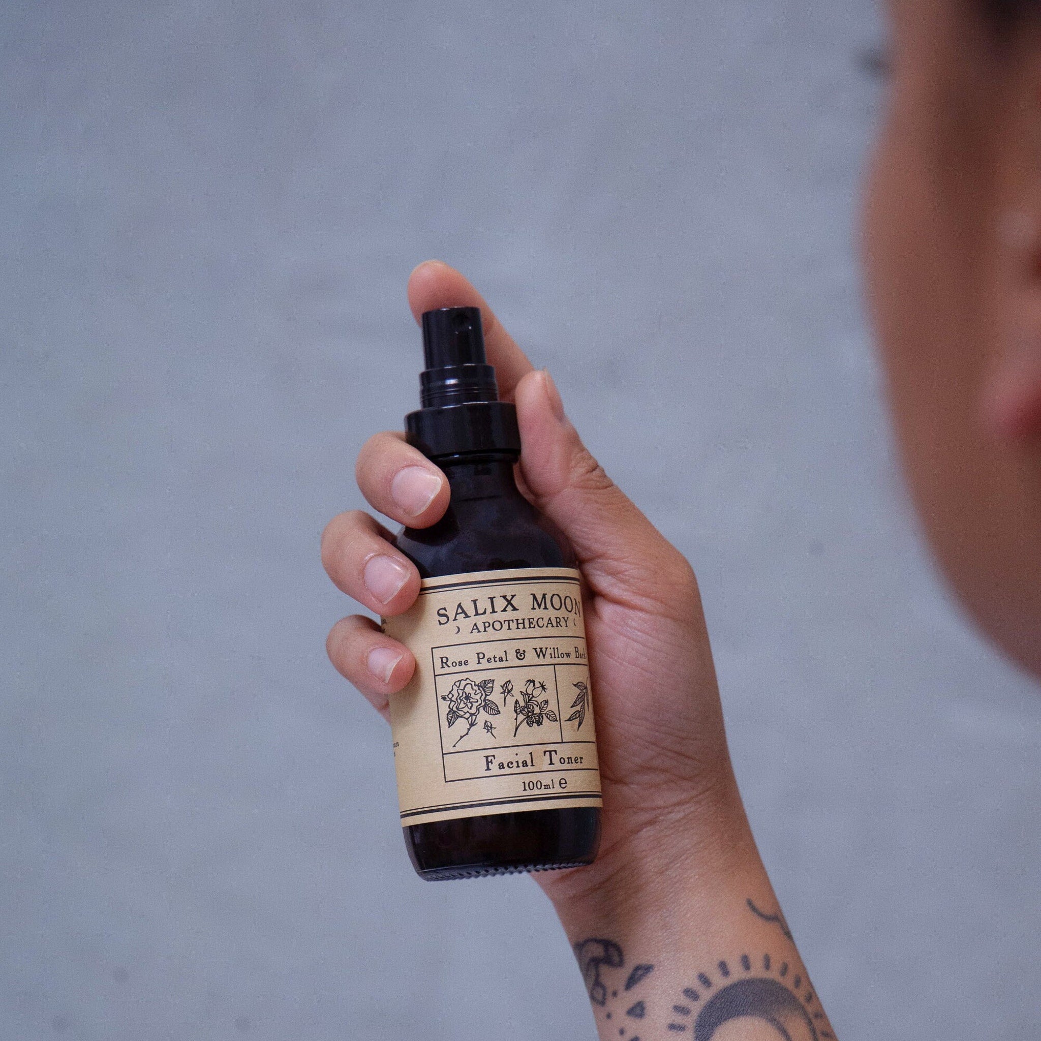 Facial toner bottle with black spray top, brown label, and held in hand
