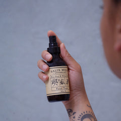 Facial toner bottle with black spray top, brown label, and held in hand