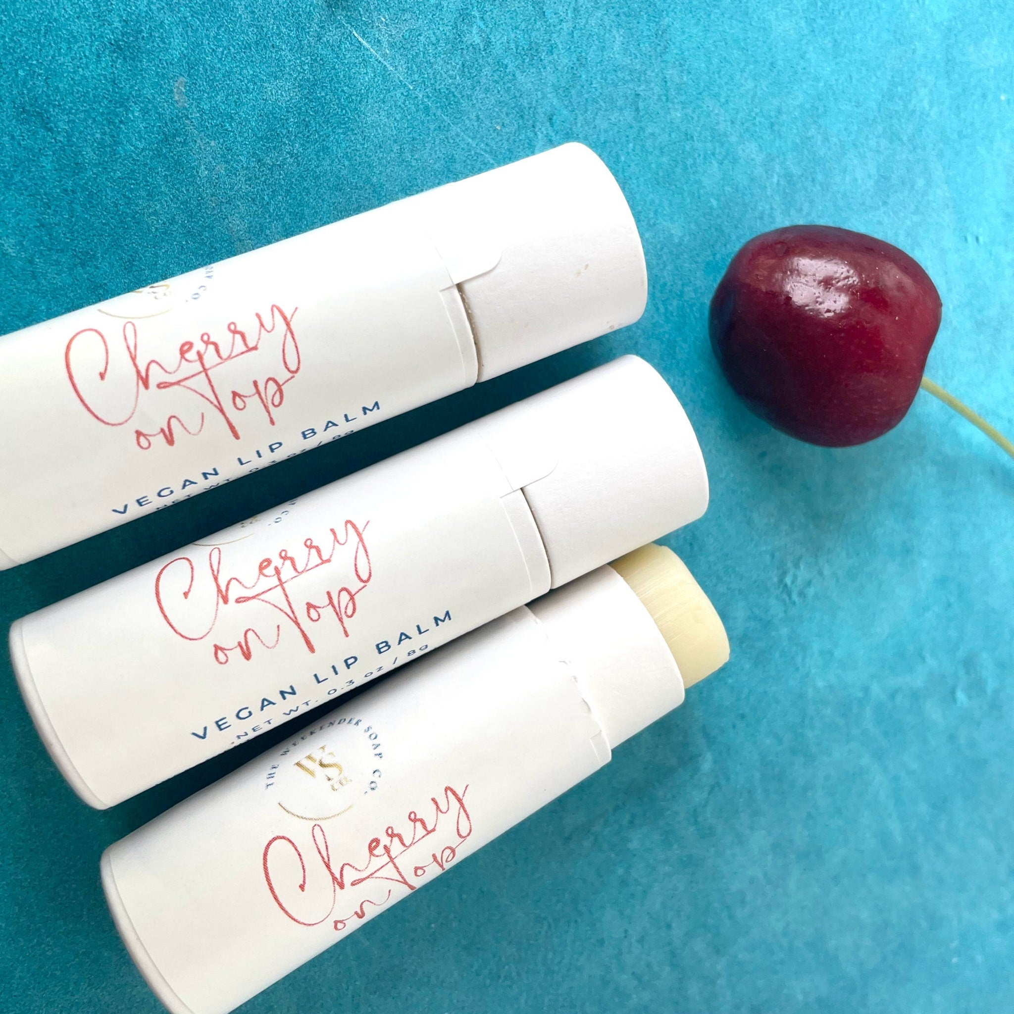 Lip balm tubes with white paper, red script text, and cherry on blue background