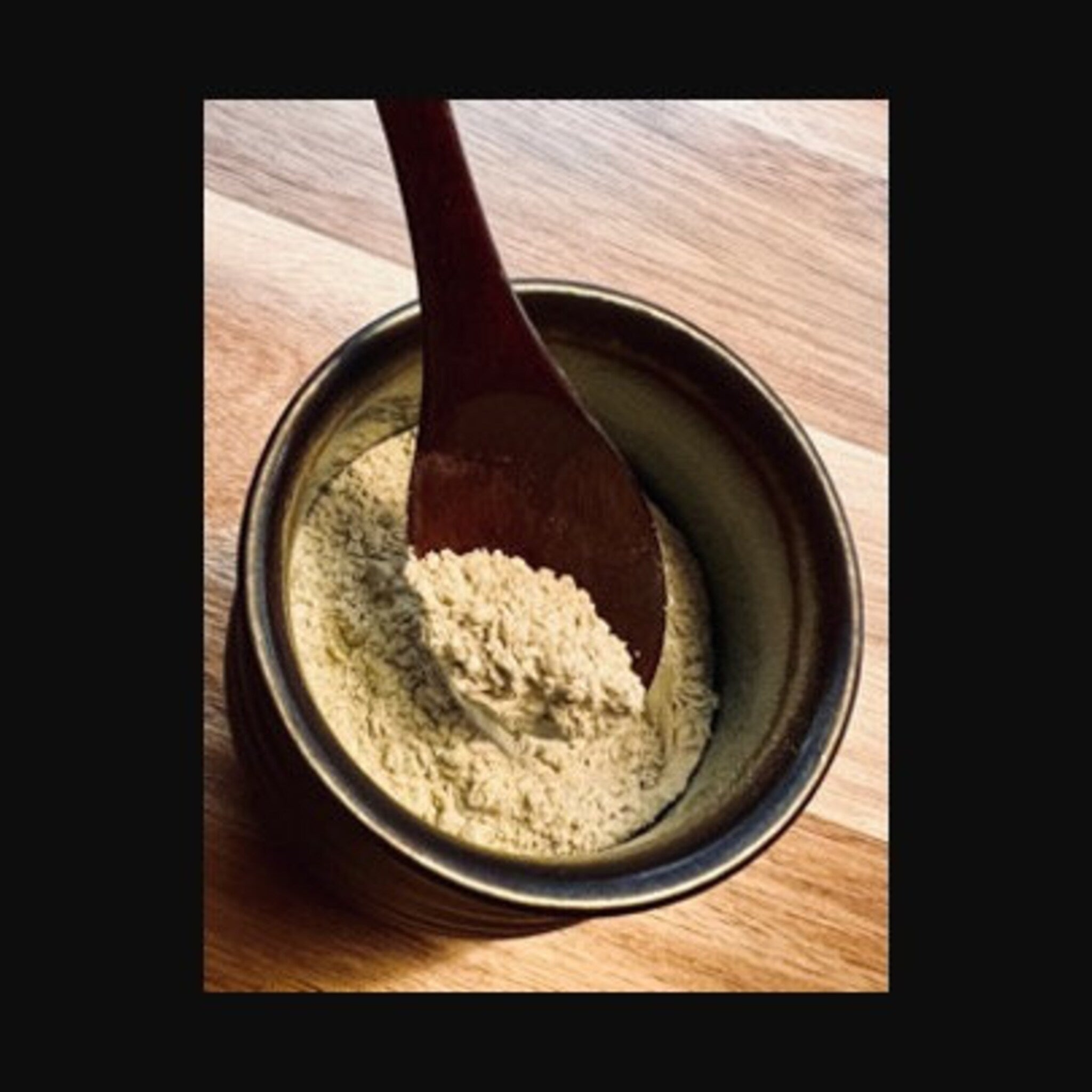 Powdered substance in a dark bowl with a wooden spoon, beige powder, dark brown spoon, wooden surface