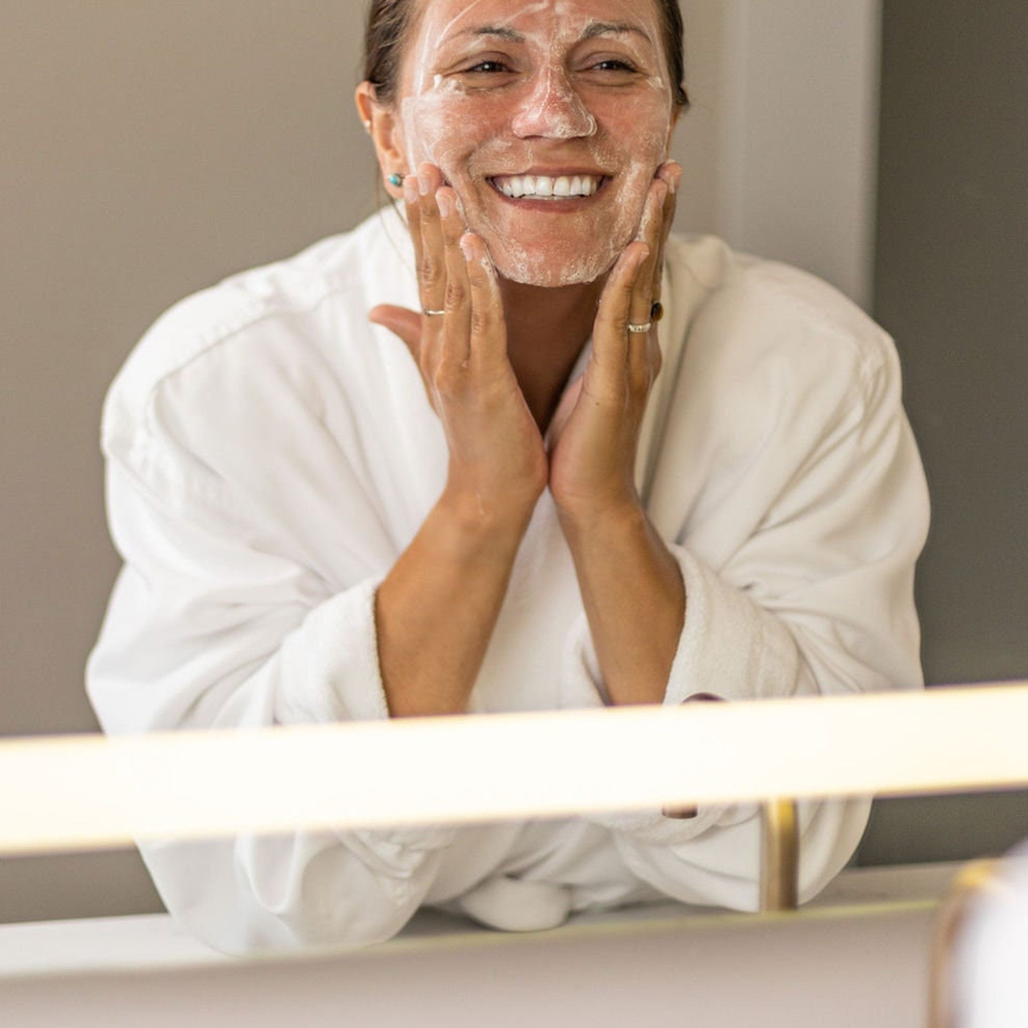 Facial cleanser application with white foam on woman's face and white robe in bathroom setting