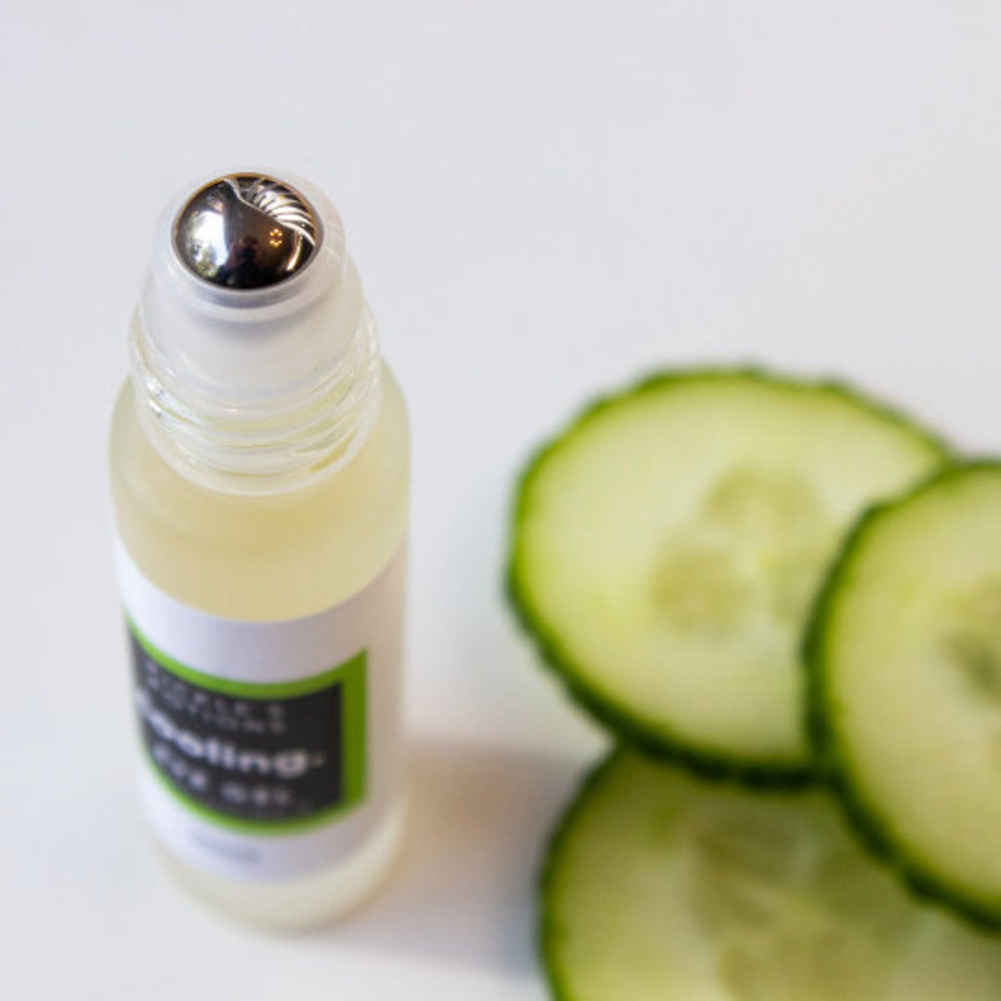 Eye oil bottle with clear glass, silver roller, and green label next to cucumber slices