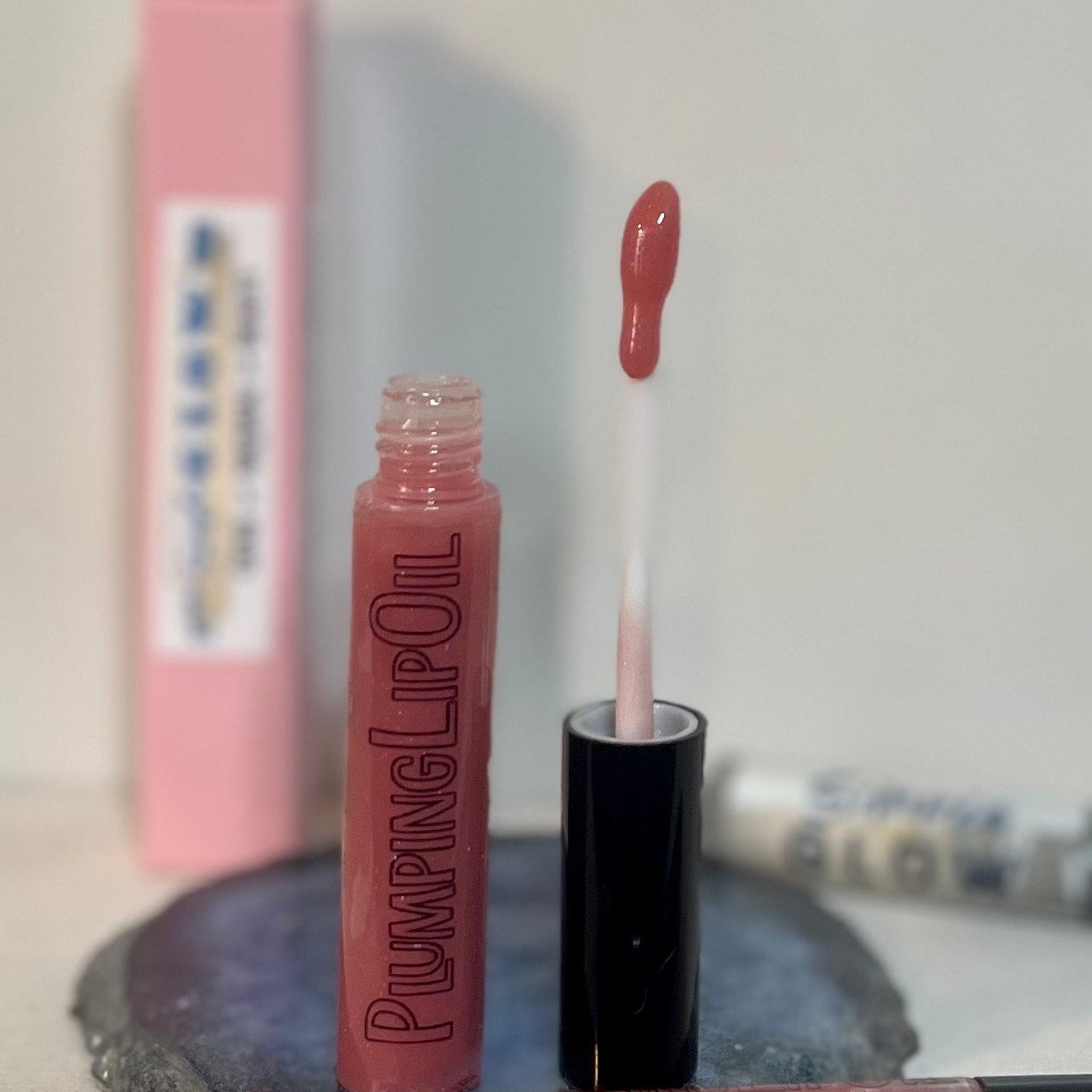 Lip oil with pink tint, clear bottle, black cap, "PLUMPING LIP OIL" text visible