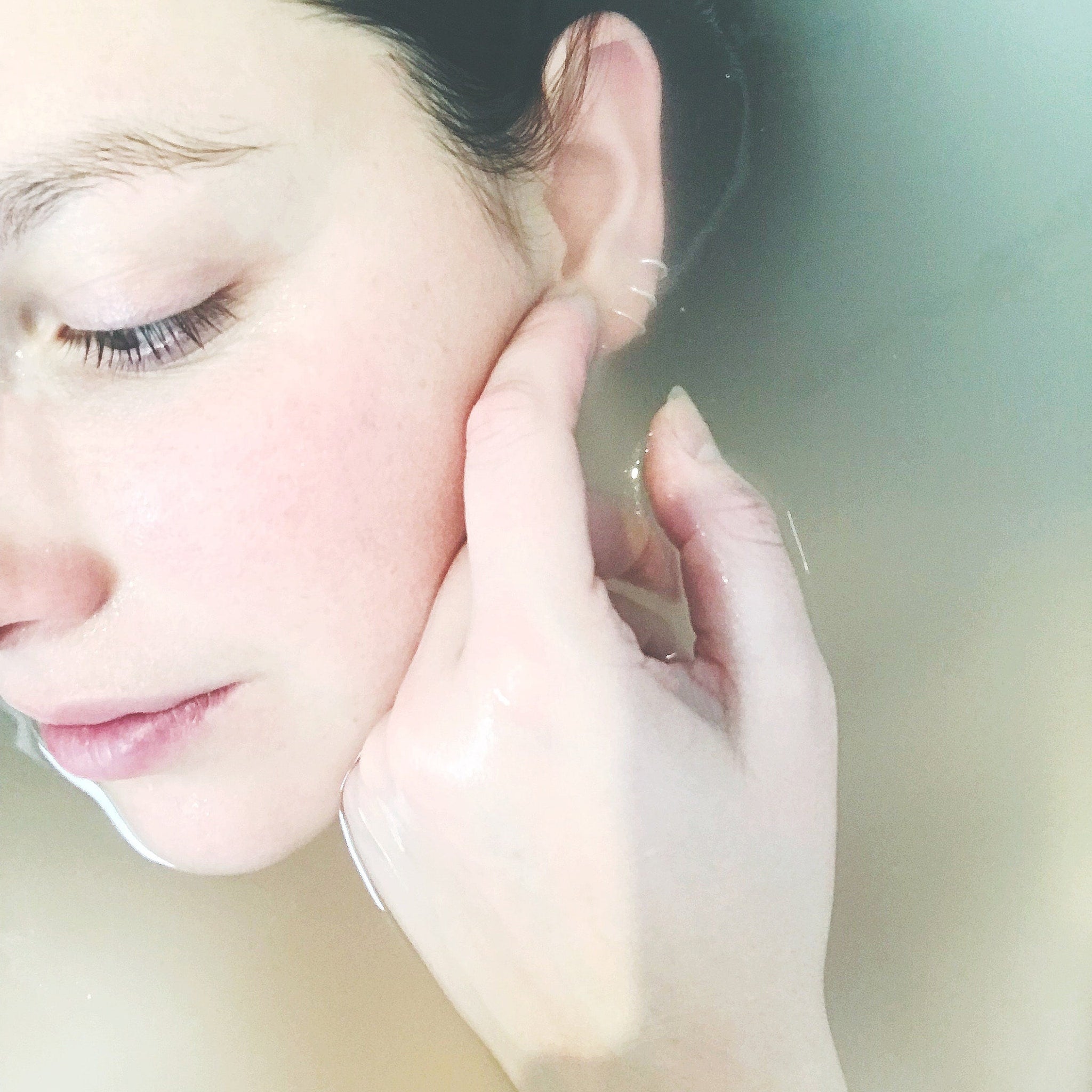 Woman's face in water, pale skin, closed eyes, hand touching ear, water around face