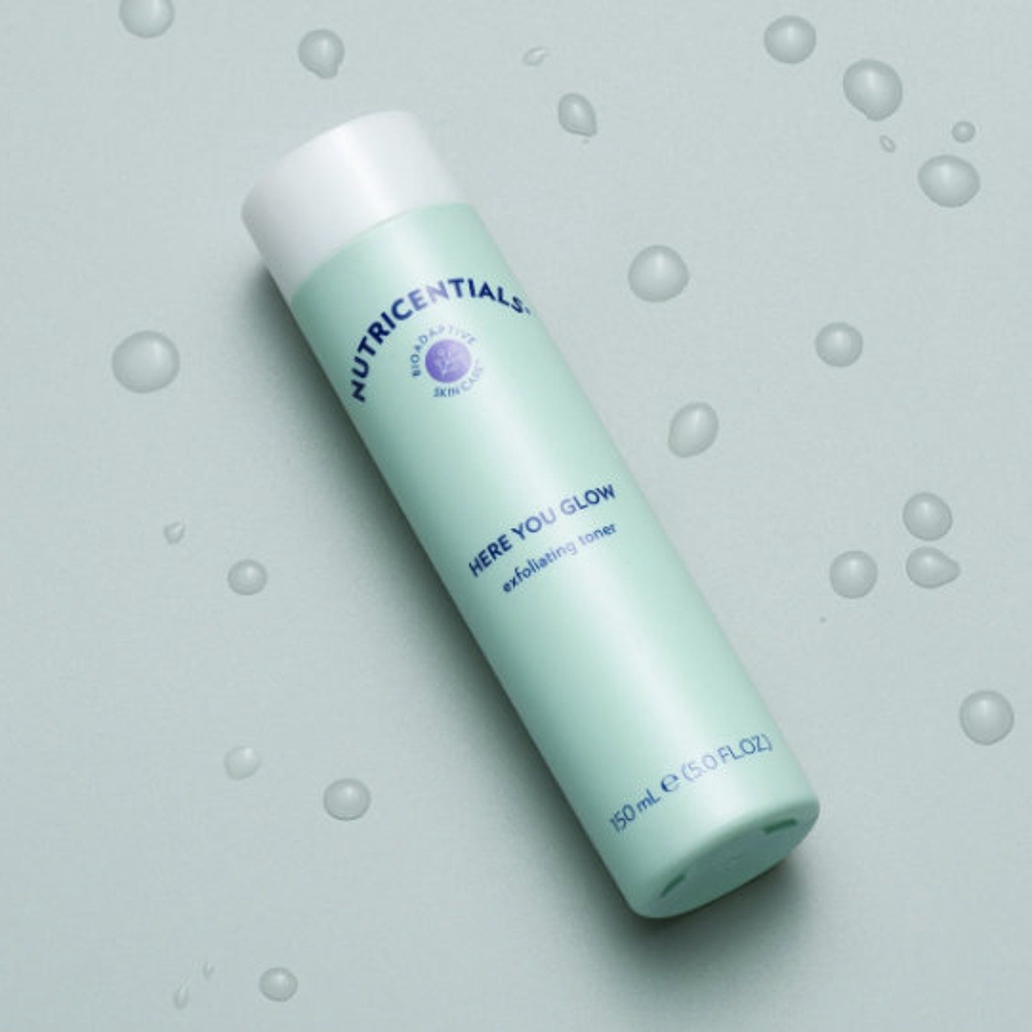 Exfoliating toner bottle, light green and white, with "Nutricentials" and "Here You Glow" text, with water droplets around it