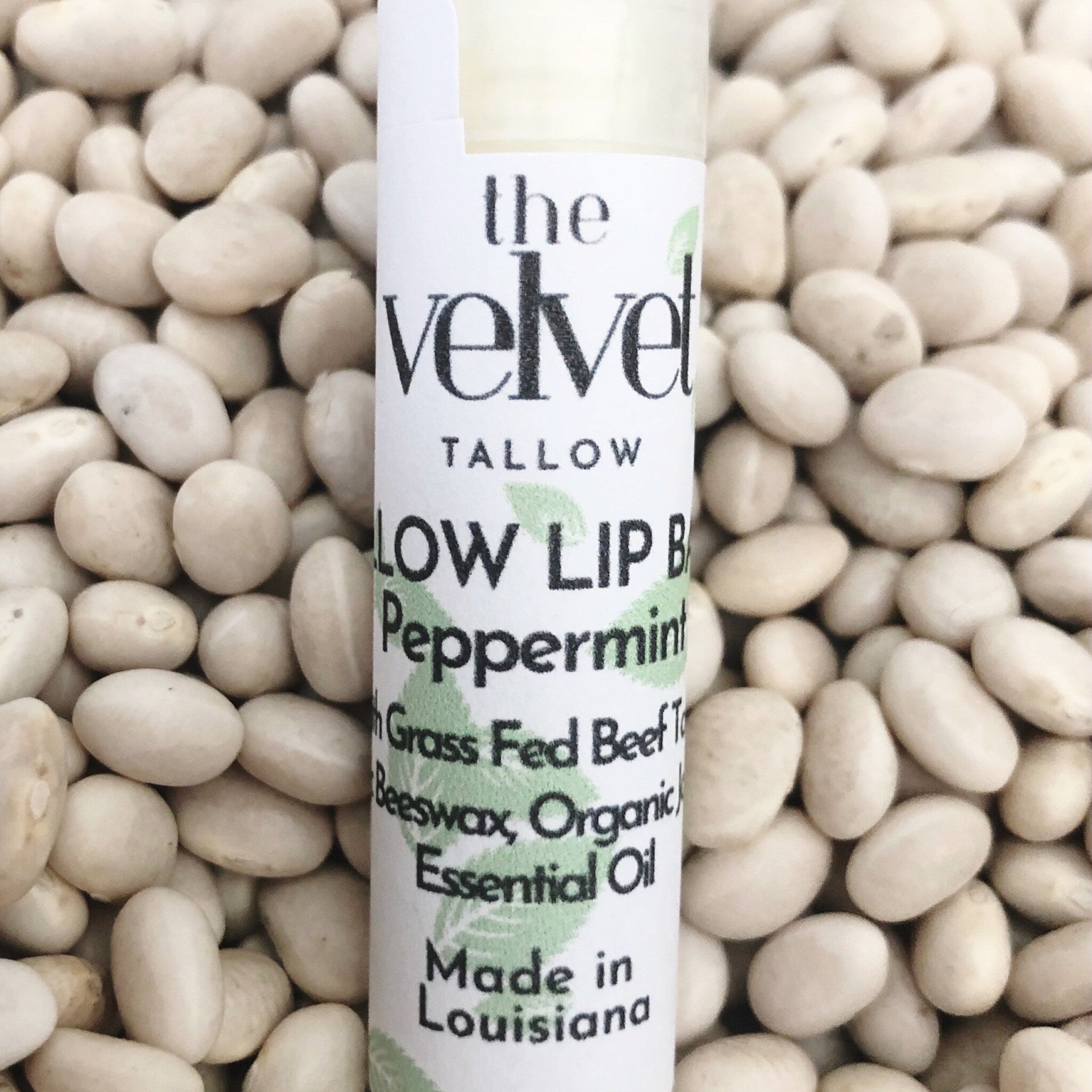 Lip balm tube with white label, black text, and white beans background. "The Velvet" brand visible.