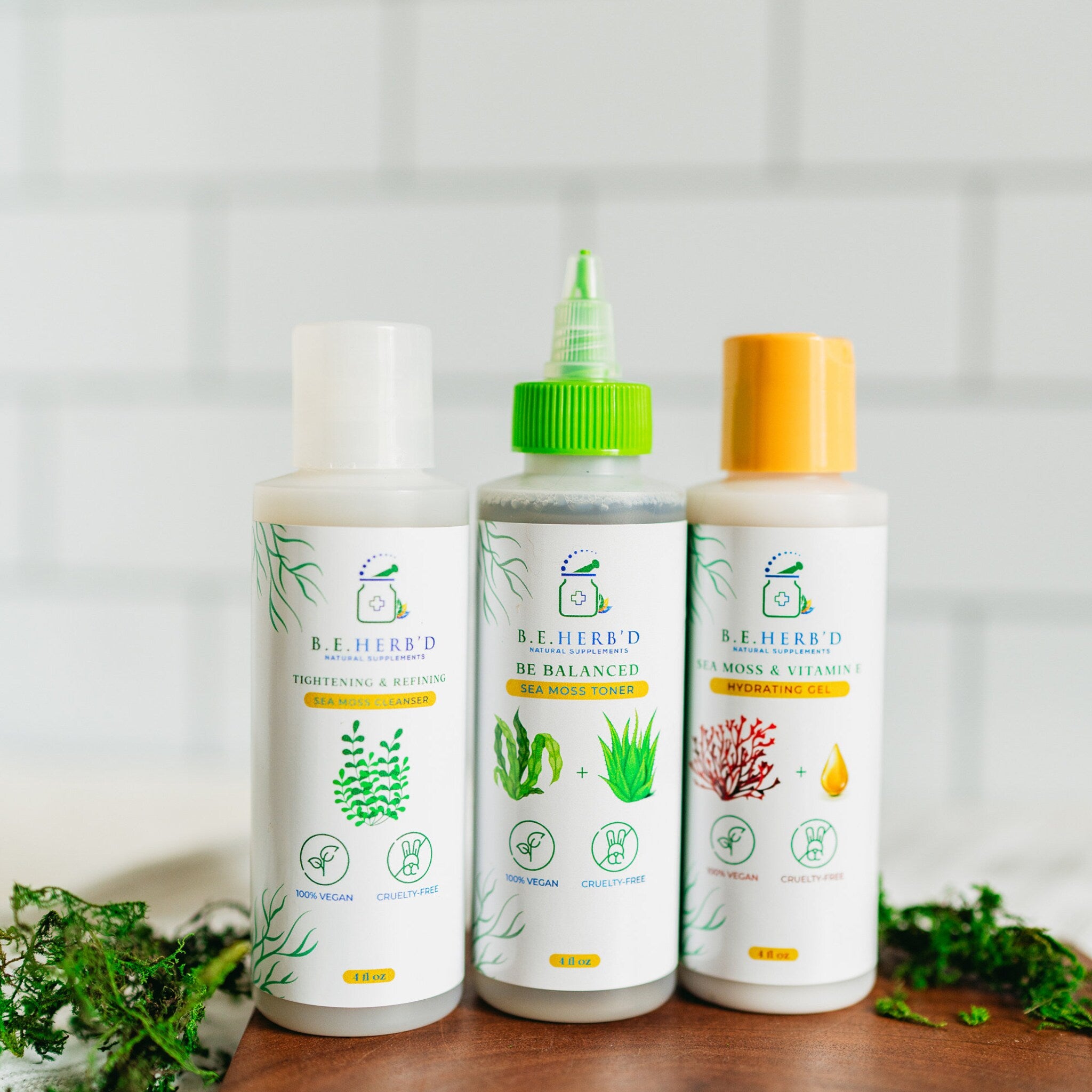 Skincare products, white bottles with green and yellow caps, "B.E.HERB'D" branding, on wooden surface