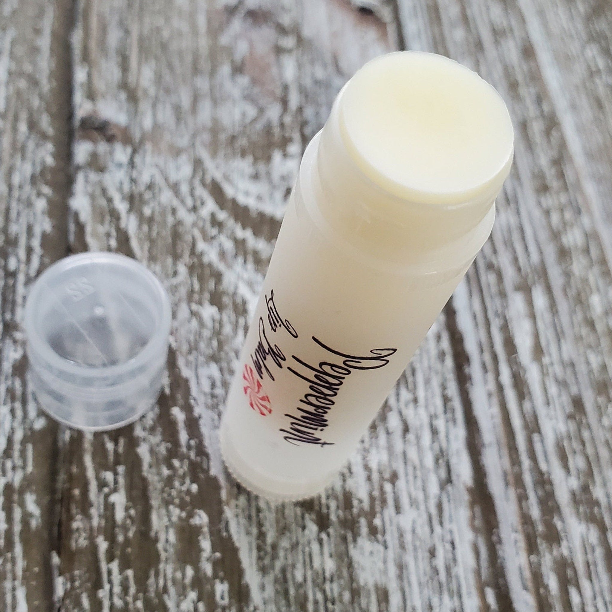 Lip balm tube with white and clear plastic, visible "Peppermint" text, and light wood background