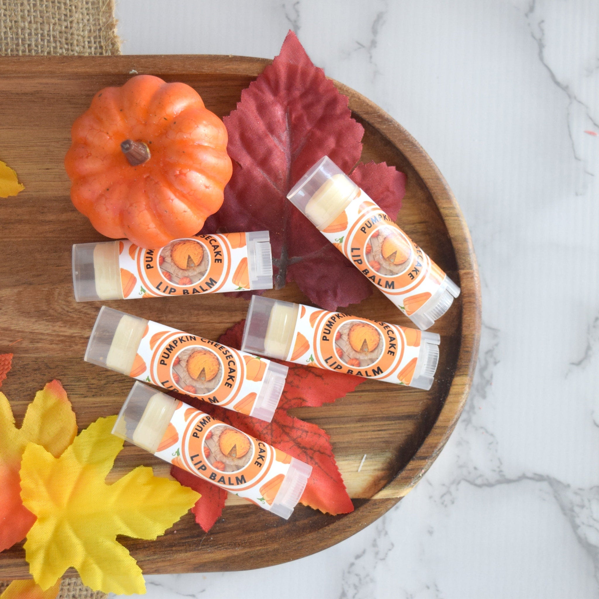 Lip balm tubes with orange and white labels, arranged with fall leaves and pumpkin on wooden tray