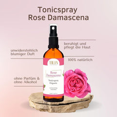 Tonic spray bottle with black spray top, amber liquid, and pink label, next to a pink rose on a rock