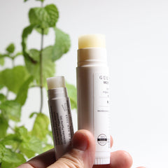 Lip balm sticks with white tubes and visible lip balm, held in hand near green leaves