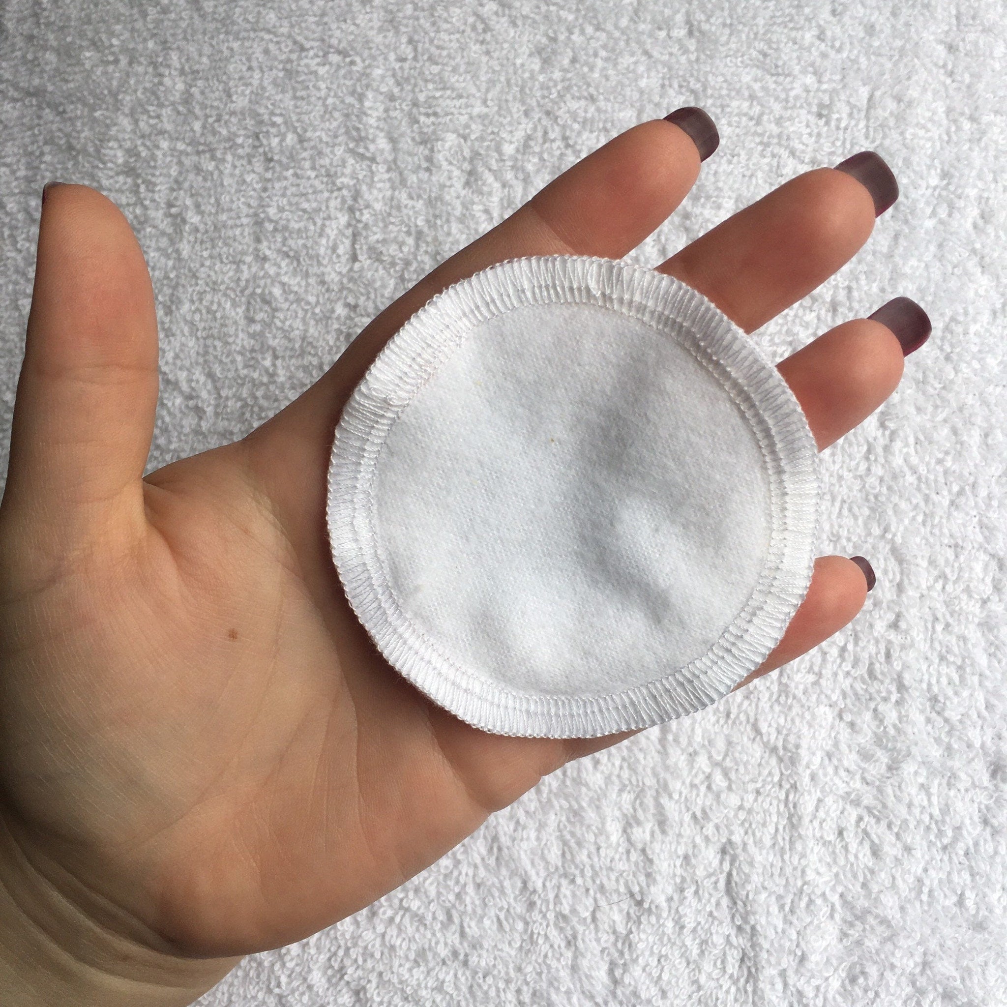 Reusable cotton pad, white fabric, stitched edge, held in hand on white towel