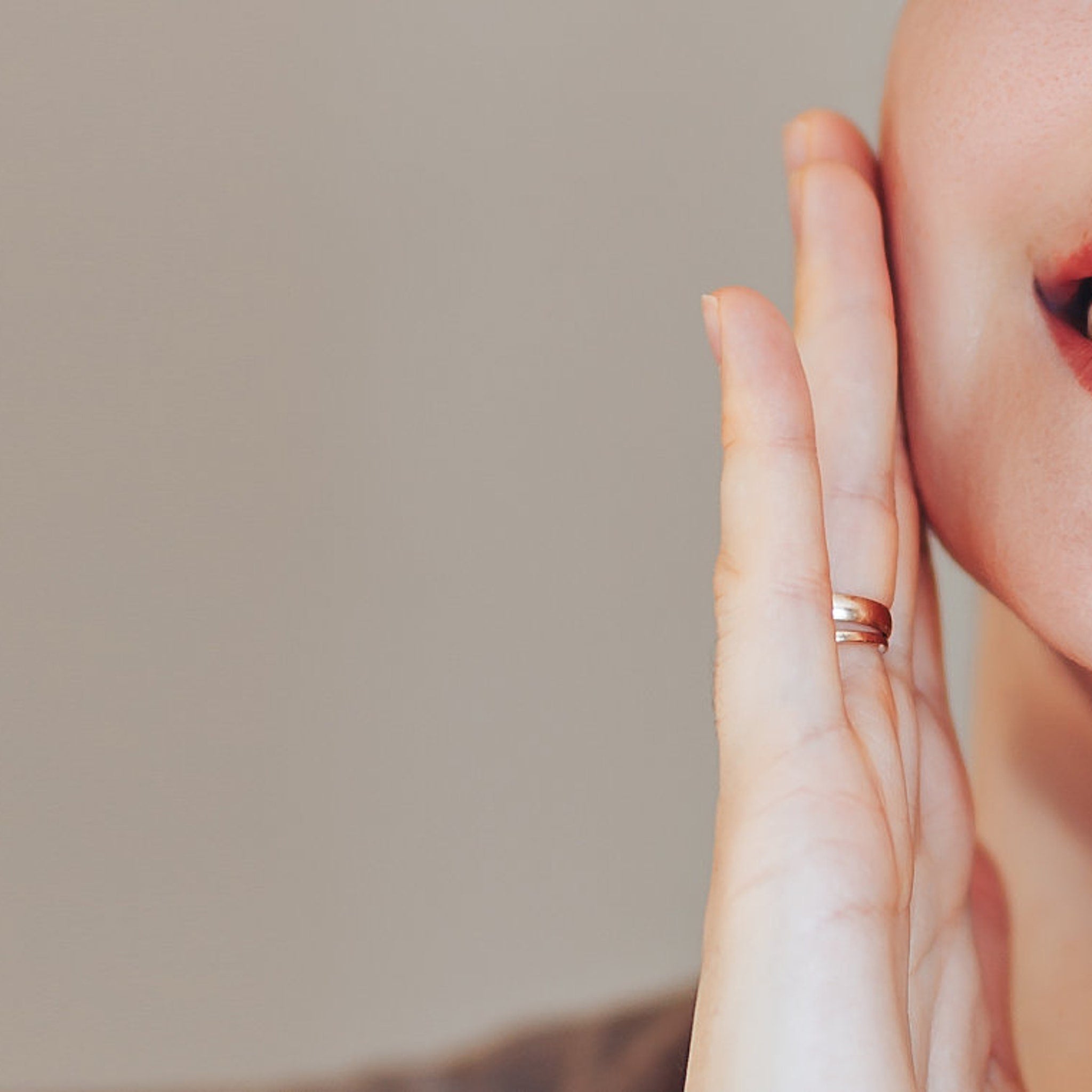 Hand with gold ring, touching face, with red lipstick and neutral background