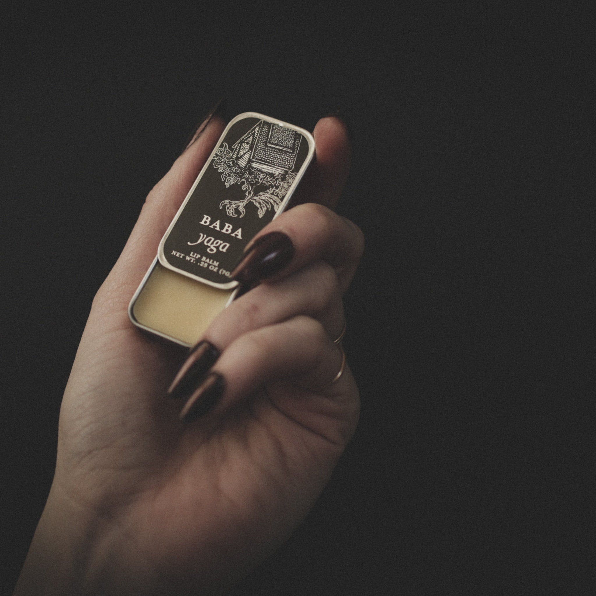 Lip balm tin held in hand, silver metal, black Baba Yaga label, yellow balm visible