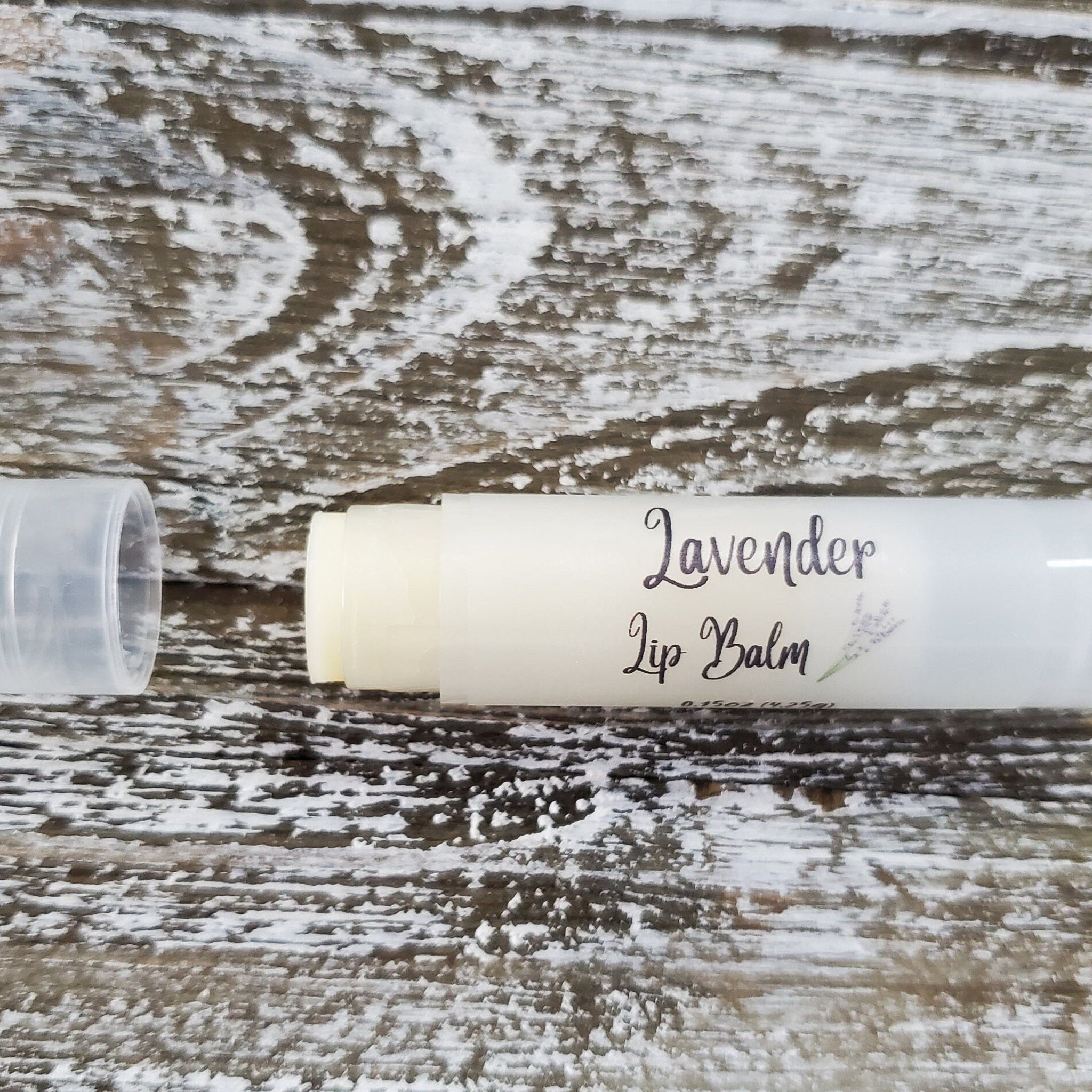 Lip balm tube with white and clear plastic, lavender text, and light purple illustration on wooden surface