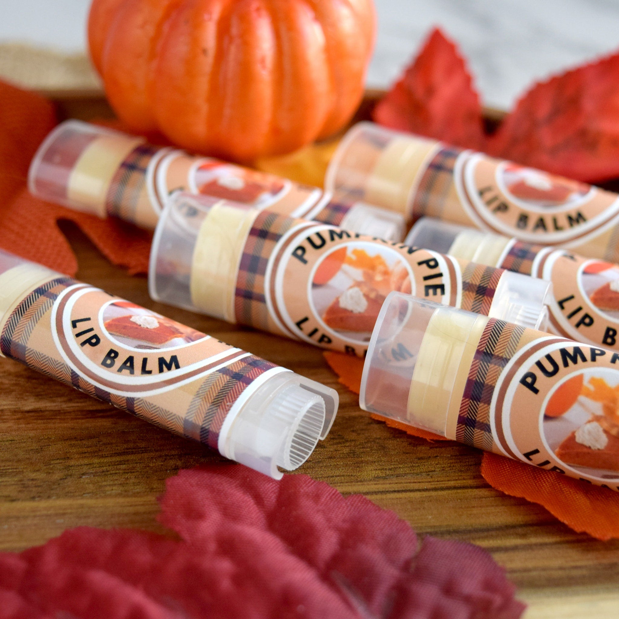 Lip balm tubes with plaid pattern and "Lip Balm" text, arranged with pumpkin and leaves