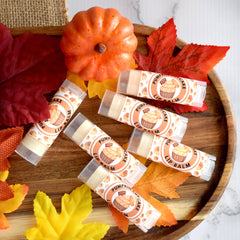 Lip balm tubes with orange and white labels, arranged on wooden tray with fall leaves and pumpkin decorations