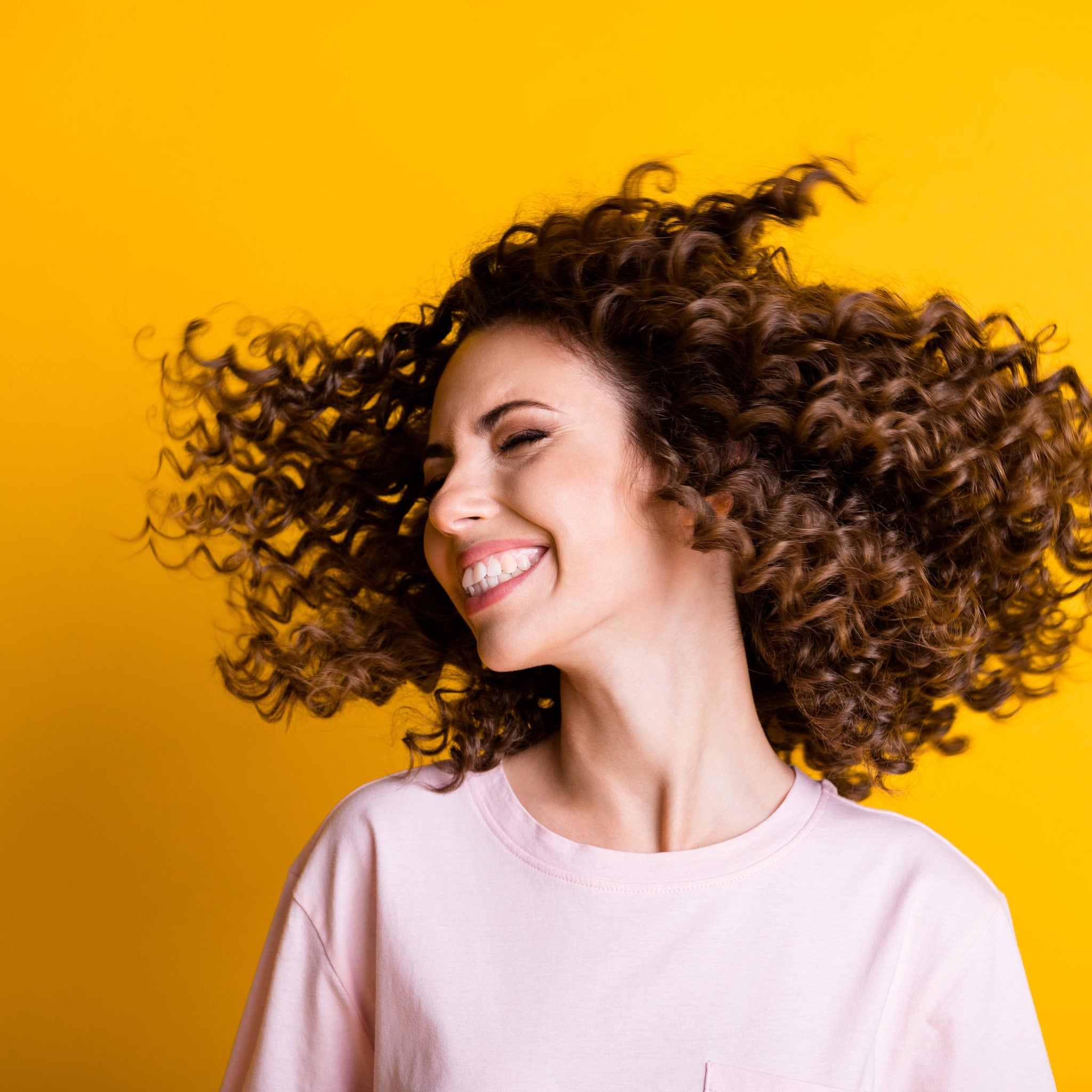 Woman with curly brown hair, wearing a pink t-shirt, smiling with eyes closed on yellow background