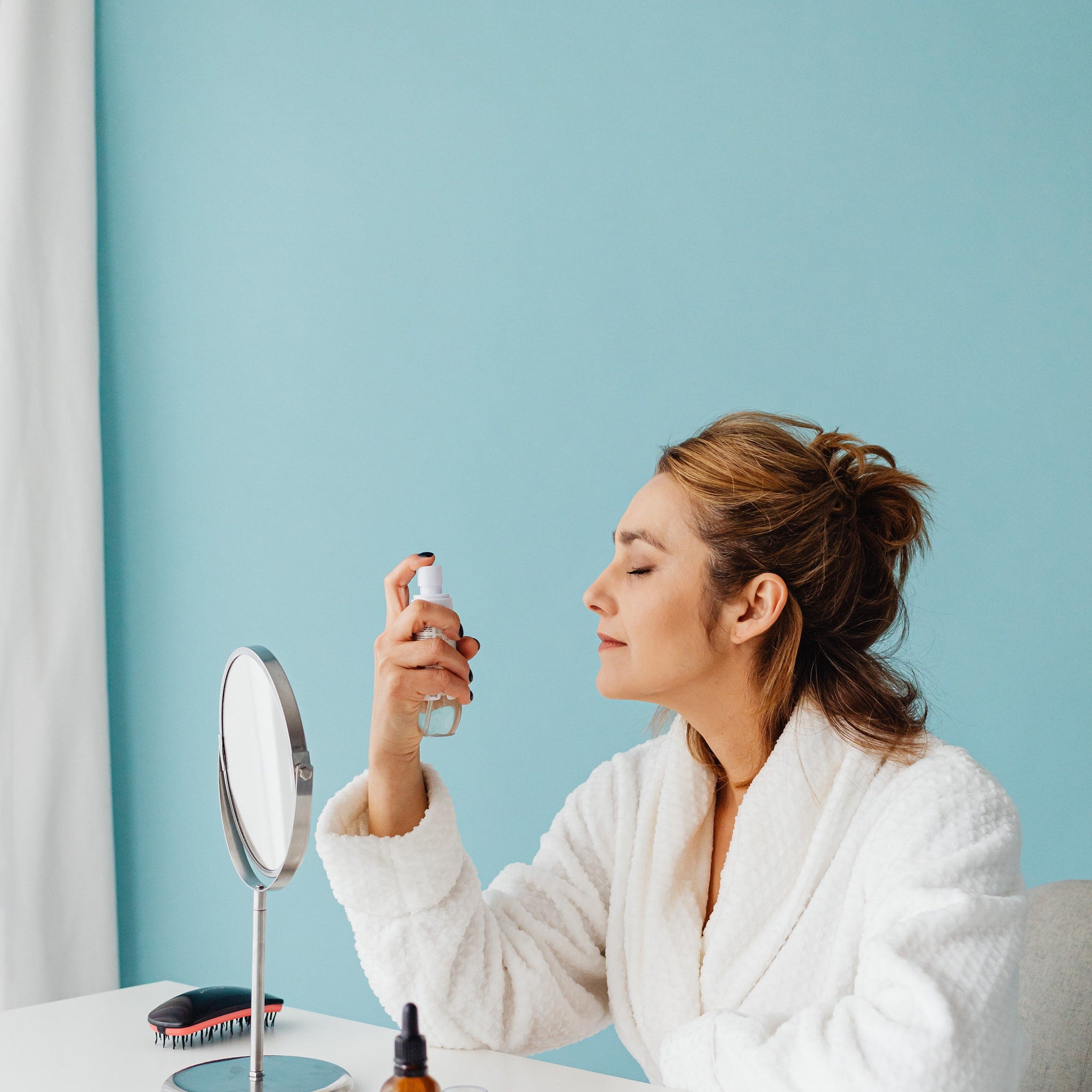 Facial mist spray bottle held by woman in white terry cloth robe near mirror