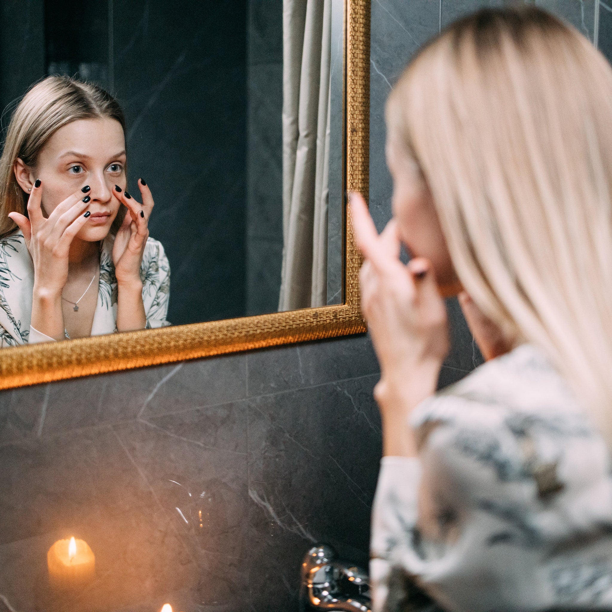 Woman looking in mirror with blonde hair and floral print robe, black nails visible