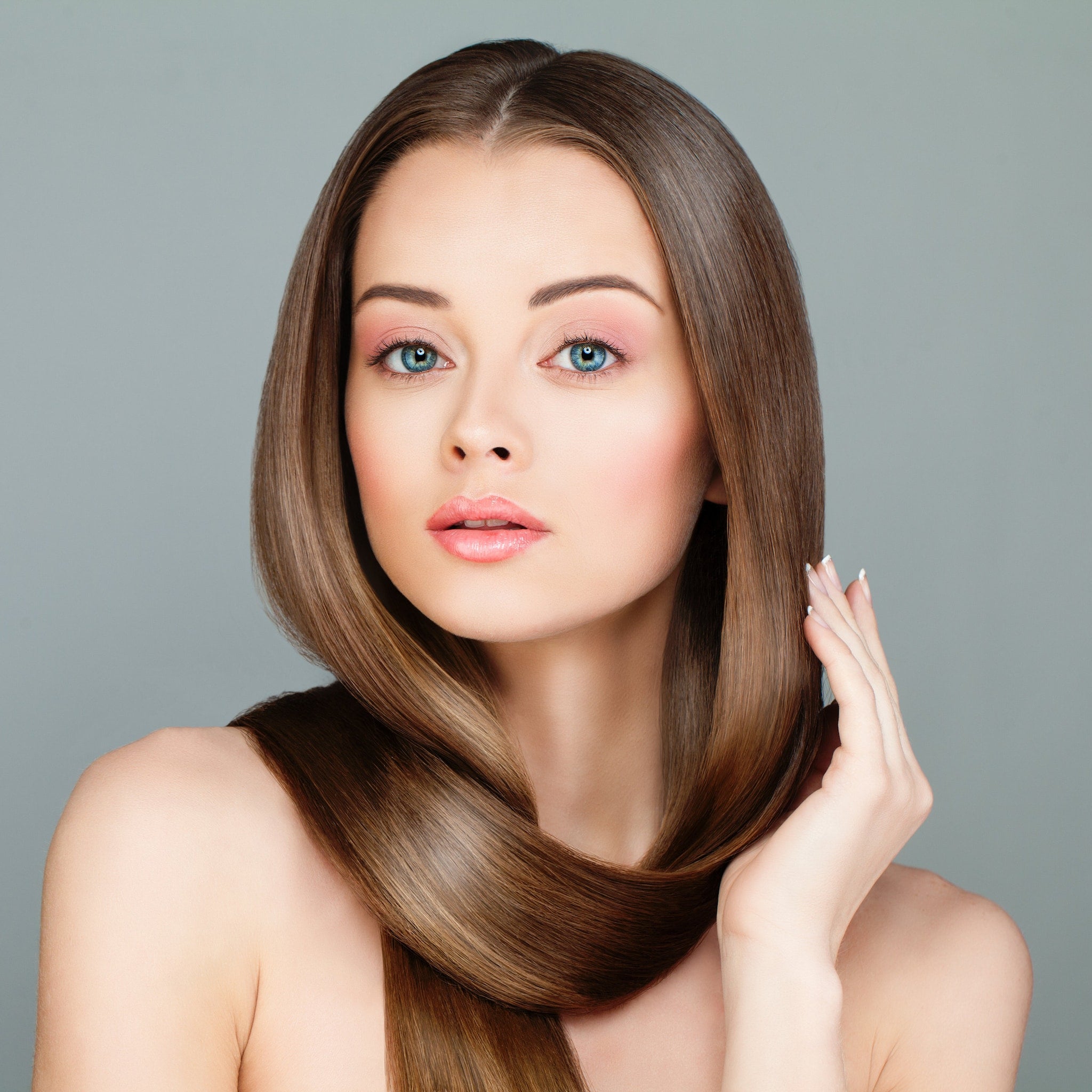 Long brown hair styled sleek, woman with blue eyes and pink makeup, holding hair
