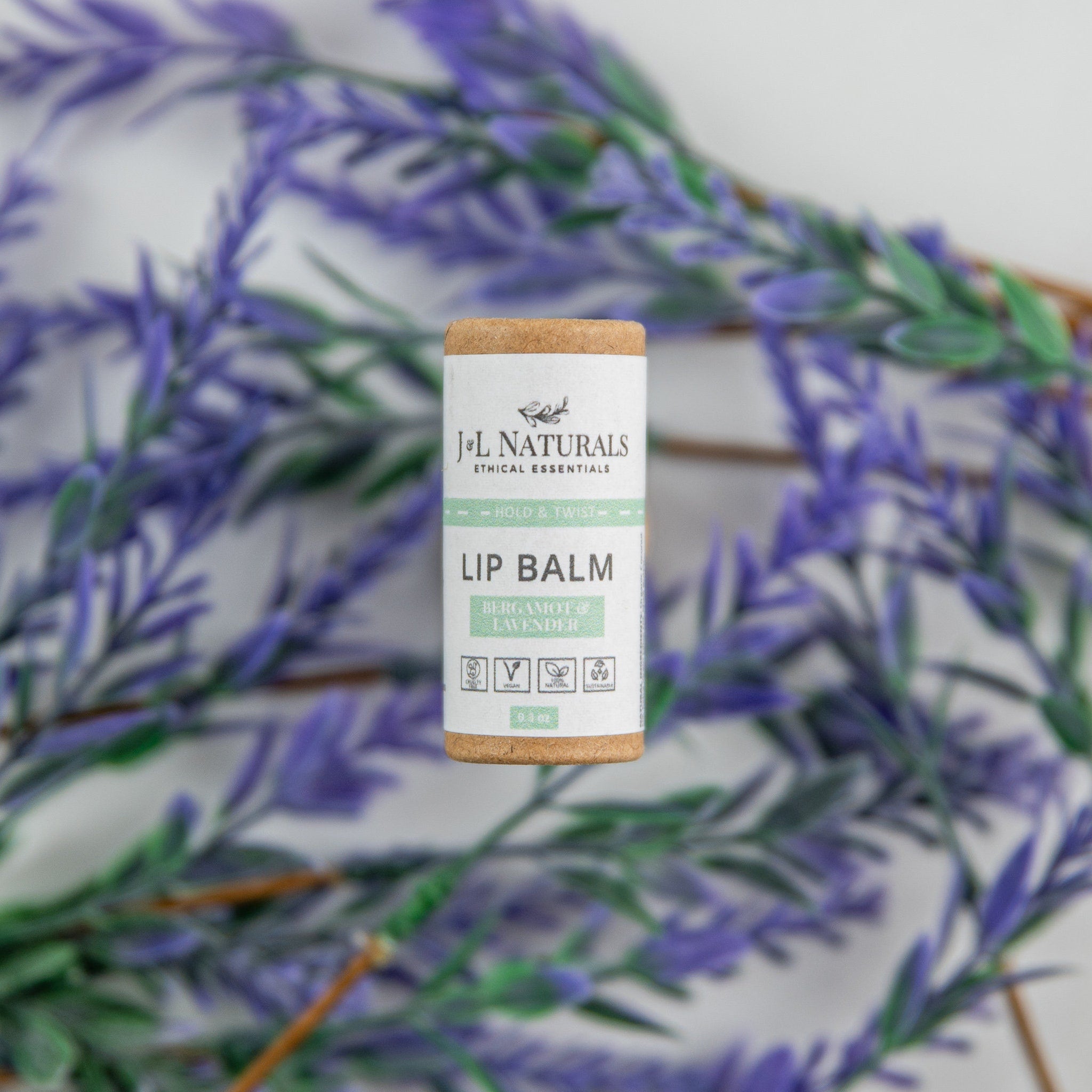 Lip balm tube with cork top, white label, and J&L Naturals branding, surrounded by purple leaves