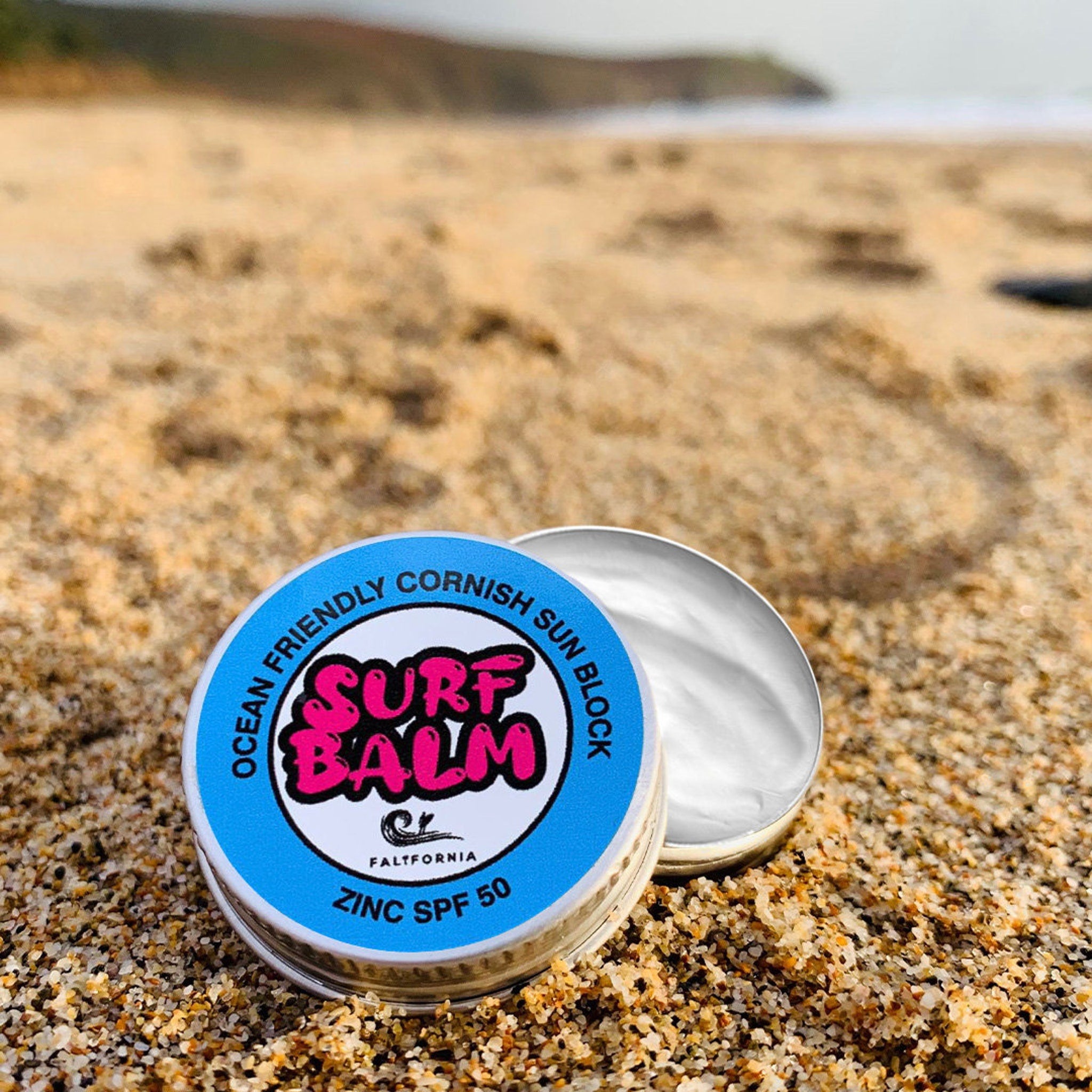 Sunscreen balm tin with blue and white label, metal container, and white cream on sand