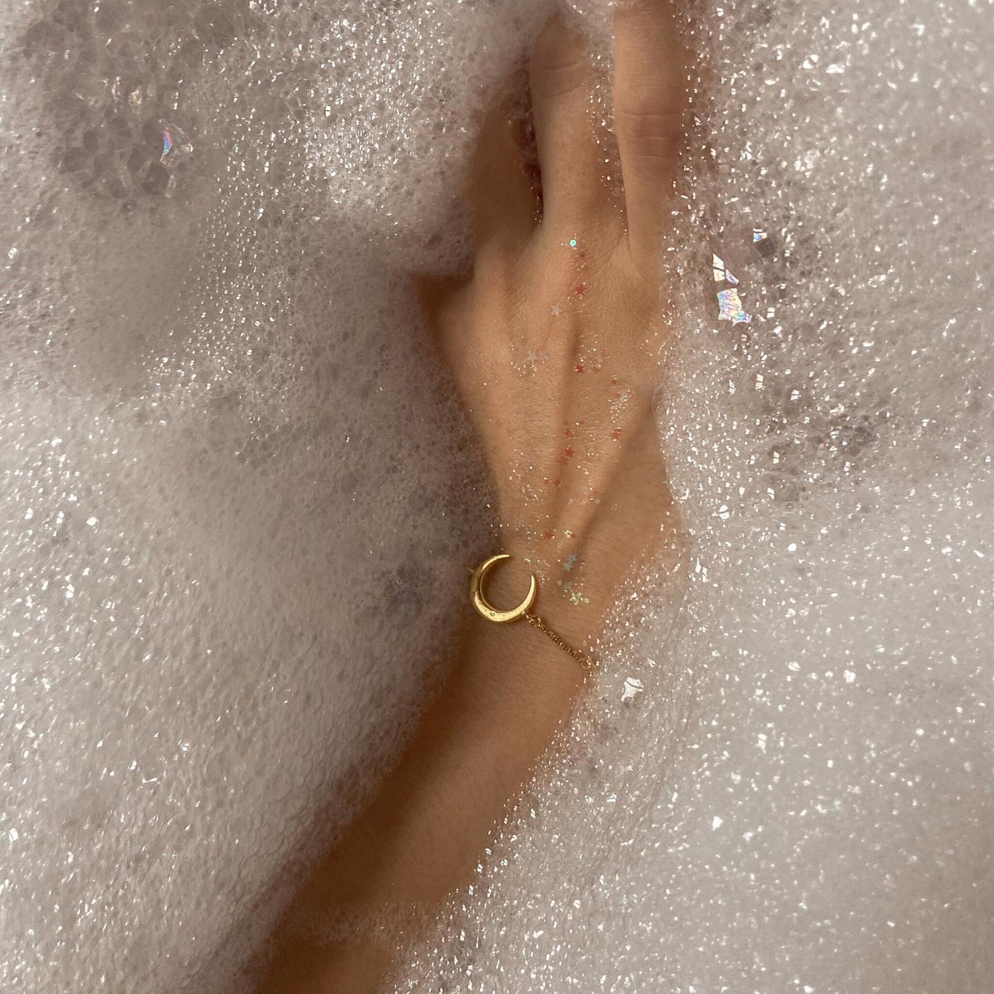 Gold crescent moon bracelet on wrist in bubbly water, gold chain detail visible
