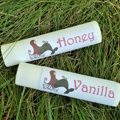 Lip balms, white tubes with red "Honey" and "Vanilla" text, Meadow Mermaids brand, on grass