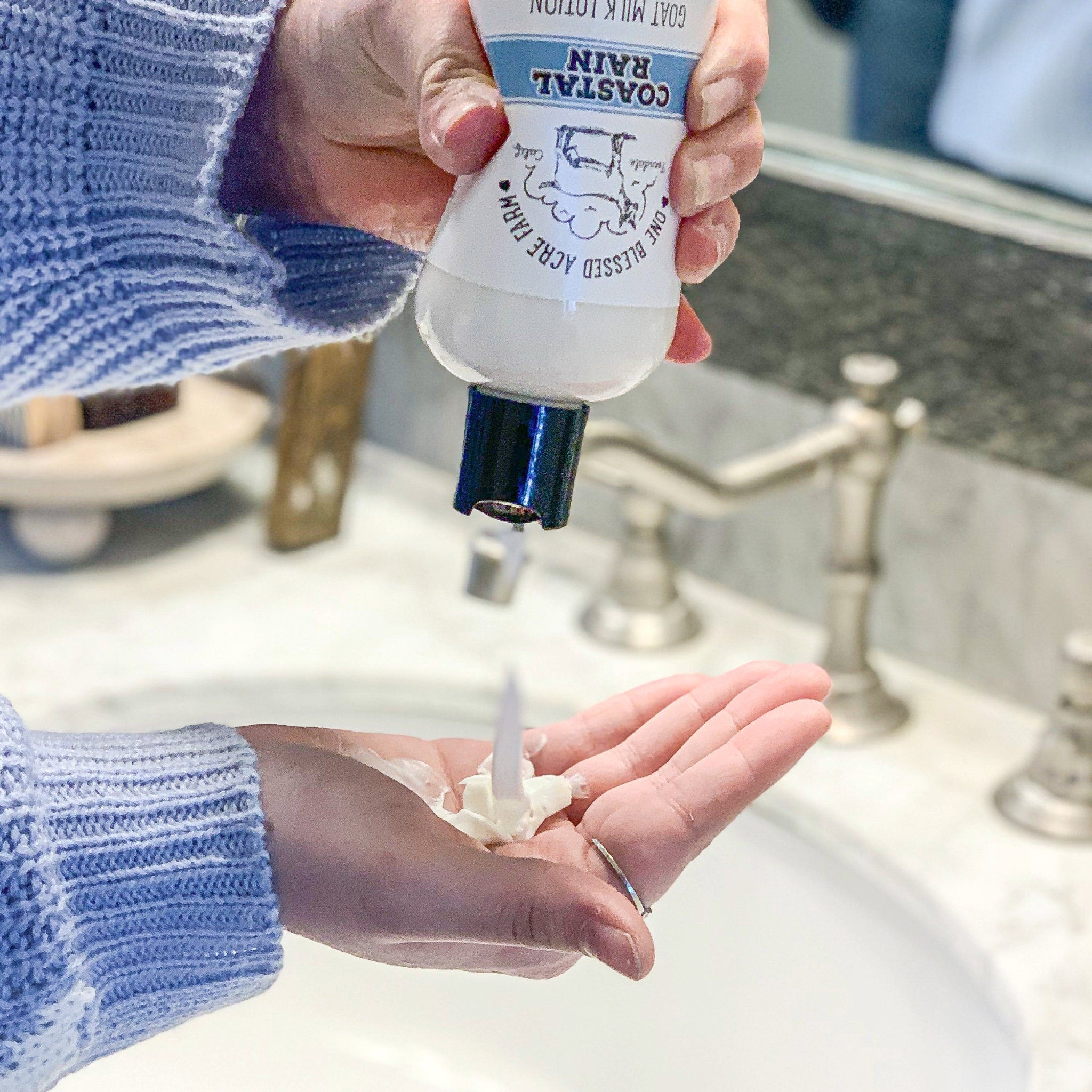 Lotion being dispensed, white lotion, blue and white Coastal Rain bottle, blue sweater sleeve, in bathroom setting