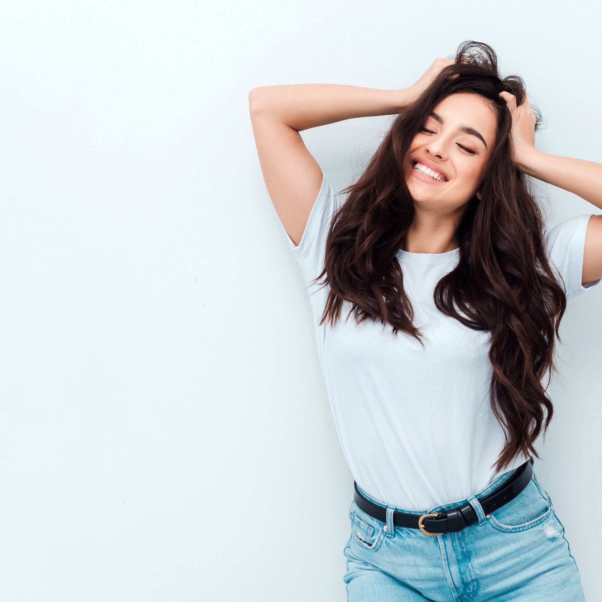 Woman wearing white t-shirt, blue jeans, black belt, and brown hair with hands in hair.