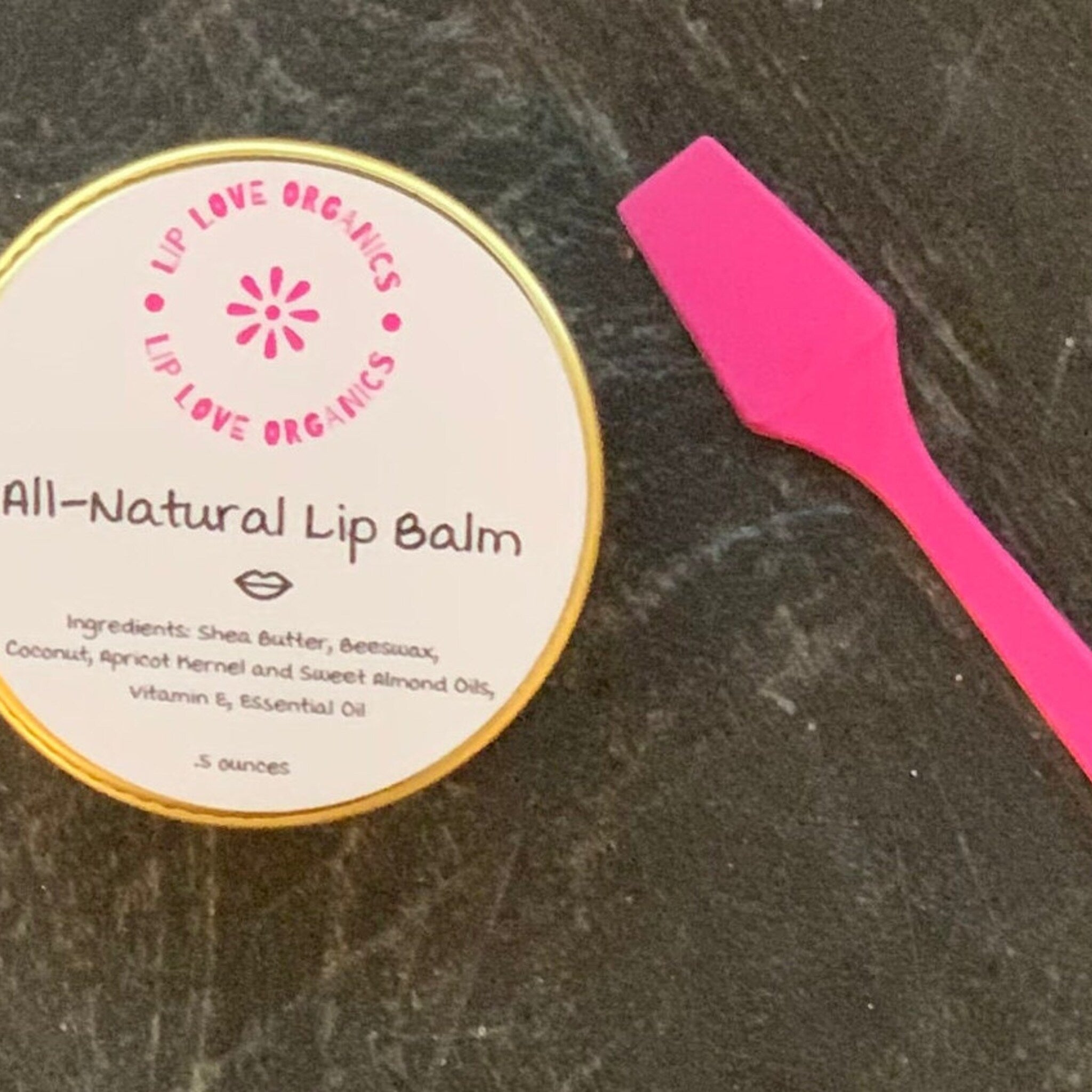 Lip balm tin with gold rim and pink applicator, "Lip Love Organics" text visible on label, on dark surface