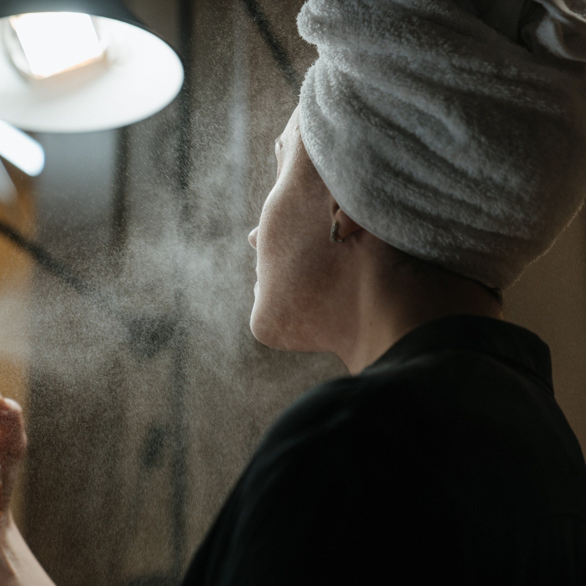 Person wearing white towel, black shirt, and being sprayed with water mist near a lamp