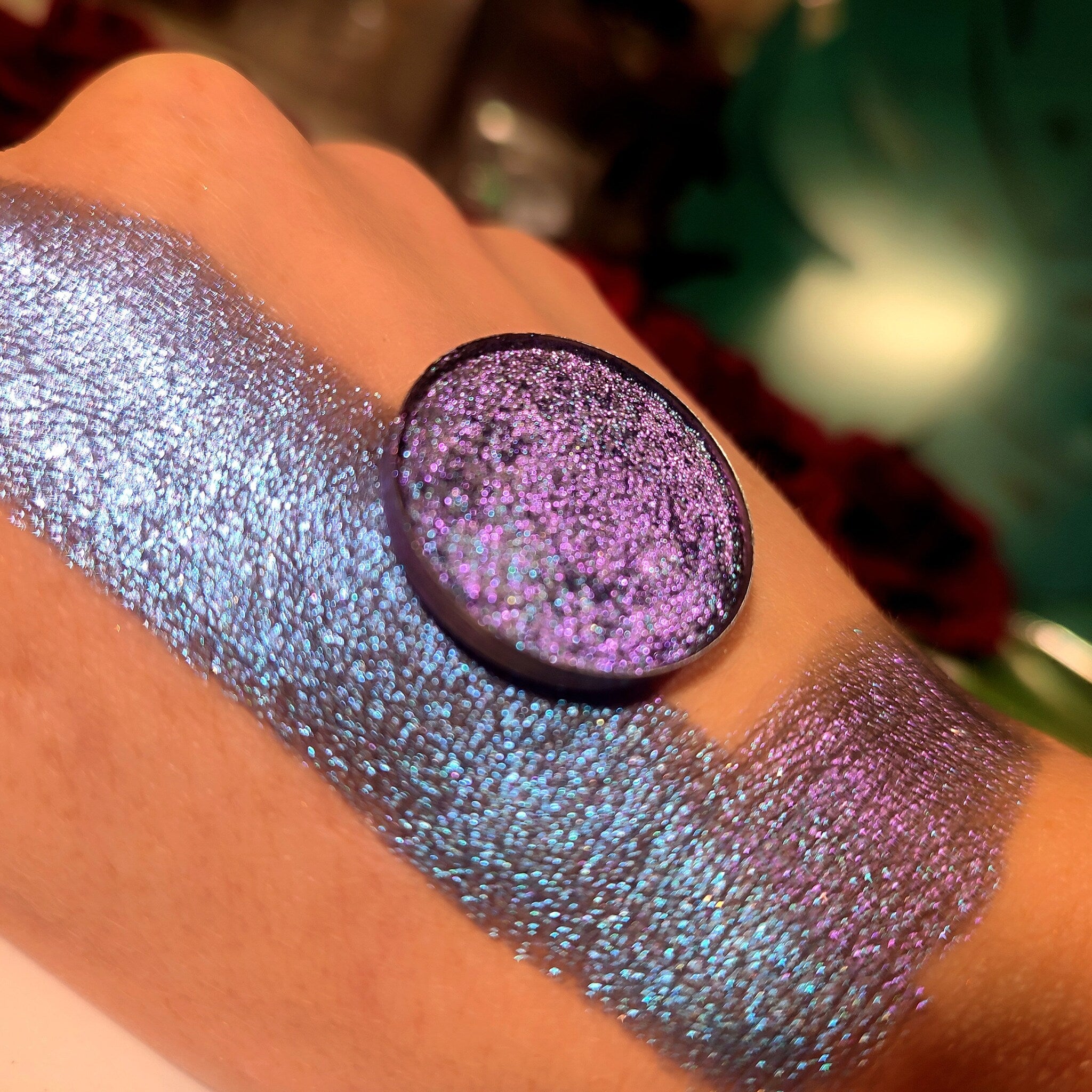 Eyeshadow swatch with iridescent purple and blue glitter on skin, round compact shown