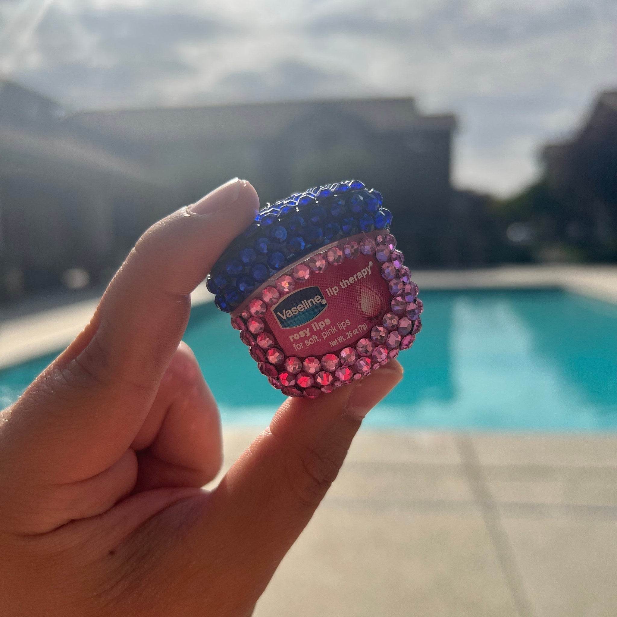 Lip therapy with blue and pink gem embellishments, held by hand with pool in background
