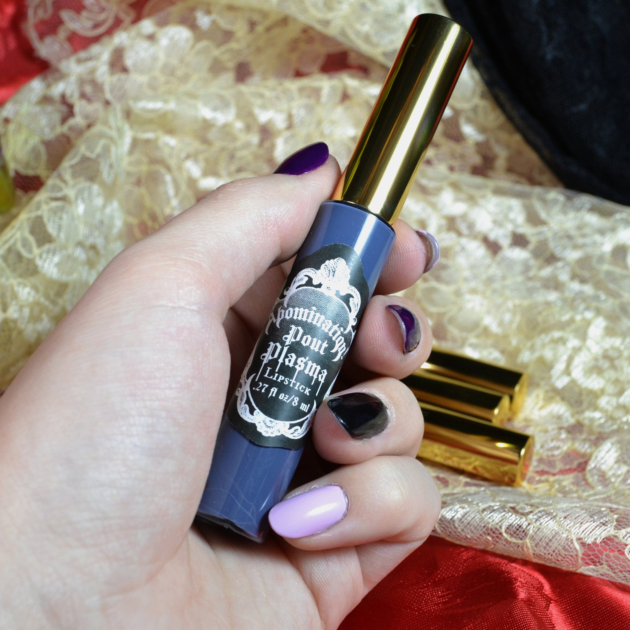 Lipstick tube in blue and gold with "Dominatrix Pout Plasma" label, held in hand with painted nails