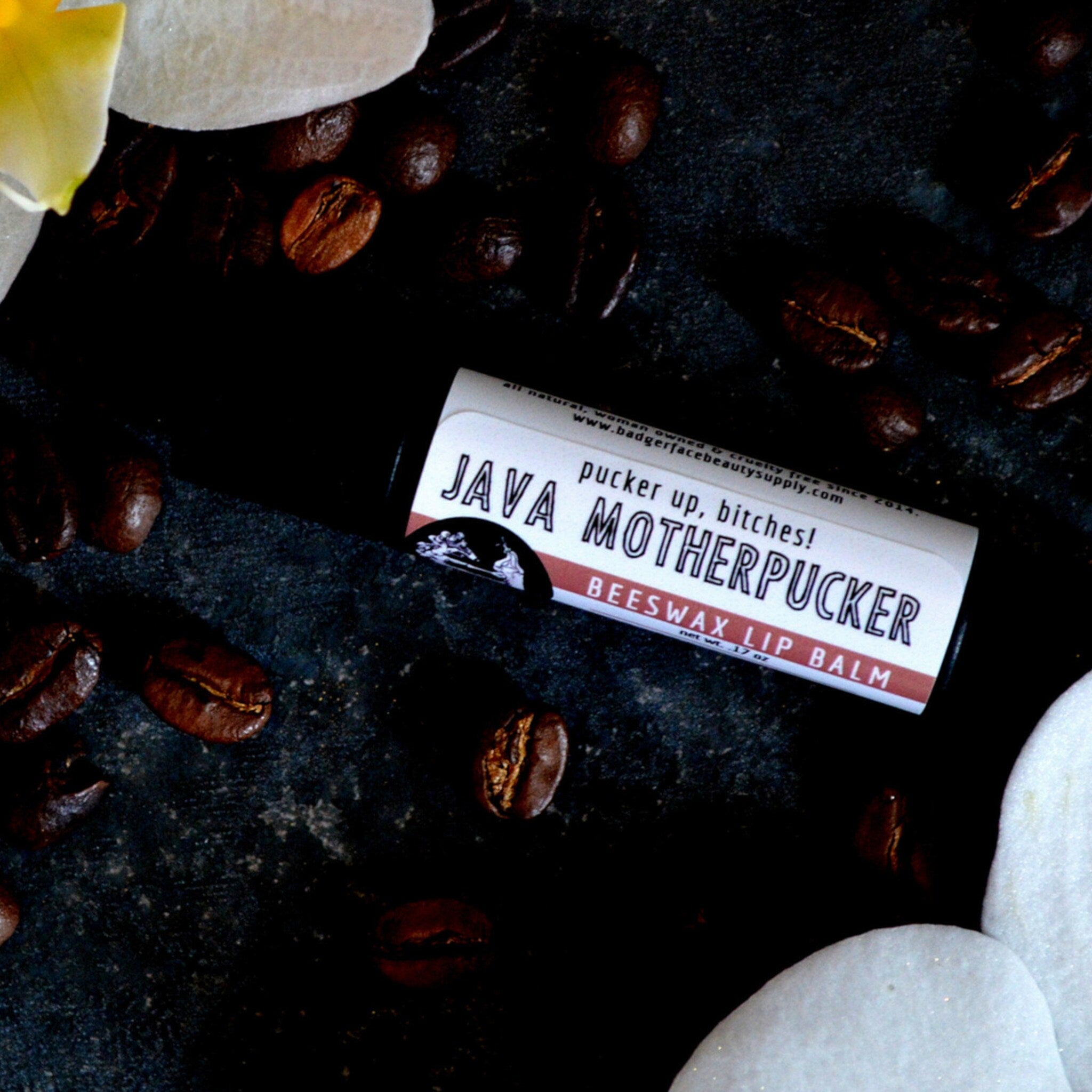 Lip balm tube with white and red label, surrounded by brown coffee beans and white flower petals, Java Motherpucker text