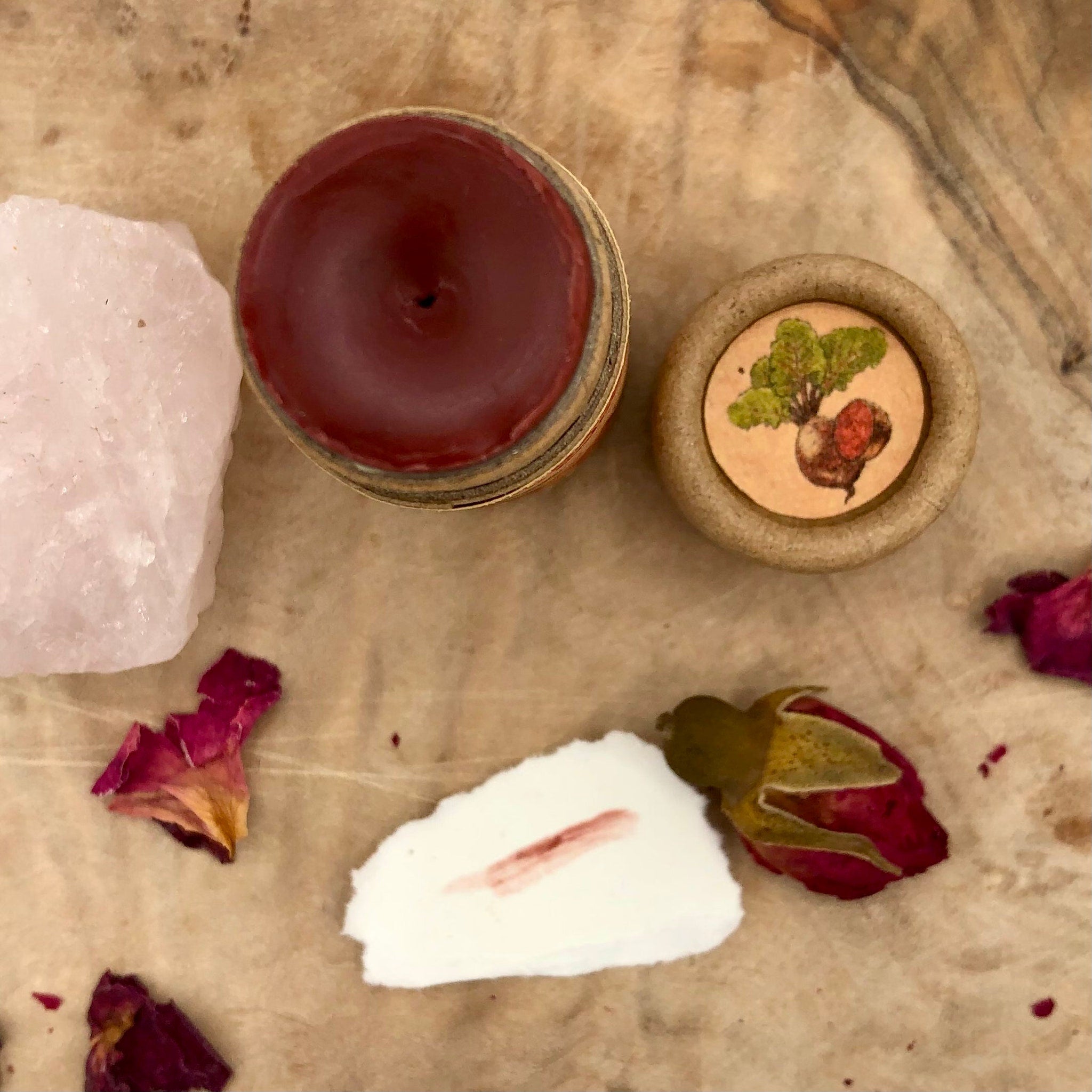 Lip balm in burgundy color, gold rim, and wooden container with beet illustration on a wooden surface