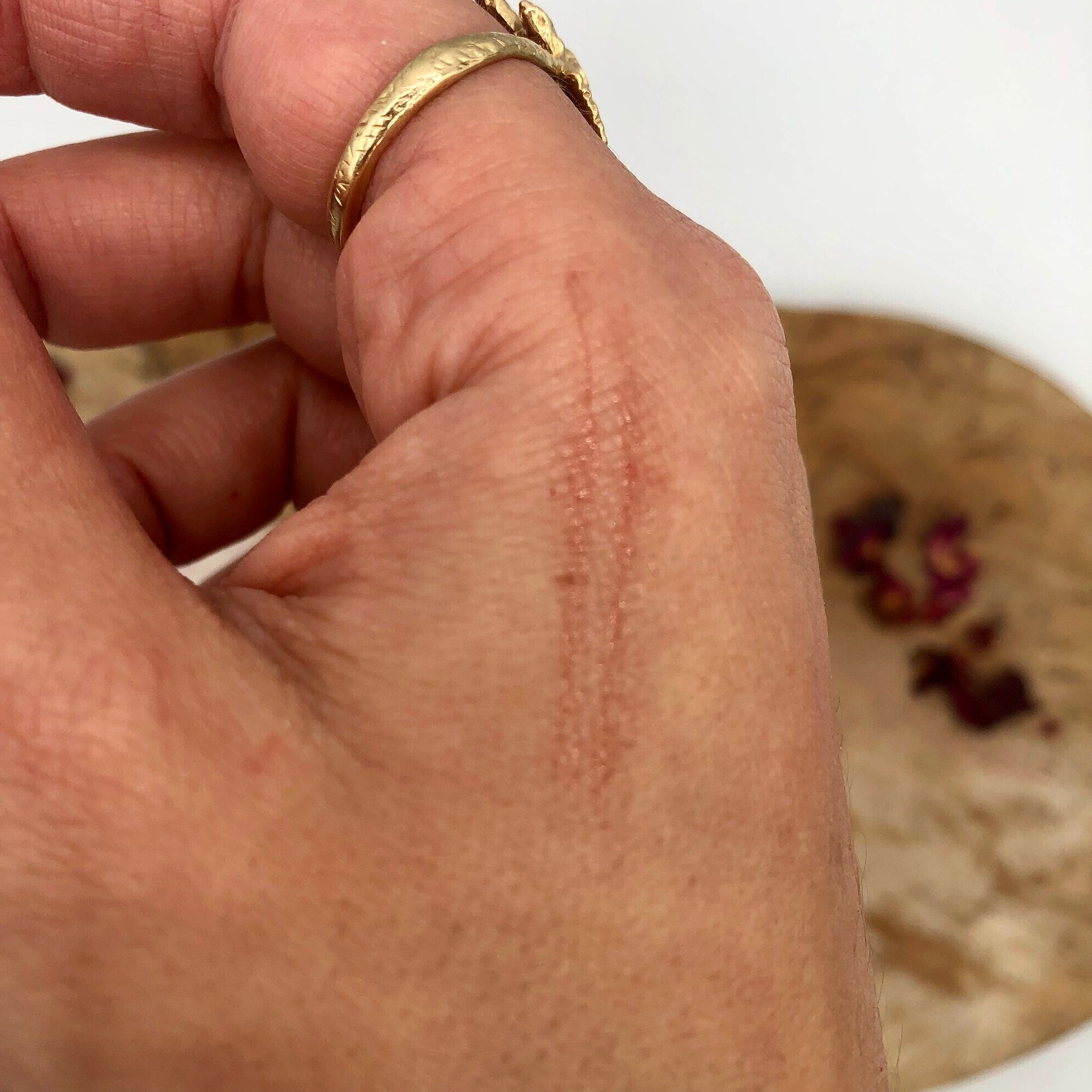 Gold ring with etched pattern on finger, resting on a wooden surface with dried flowers